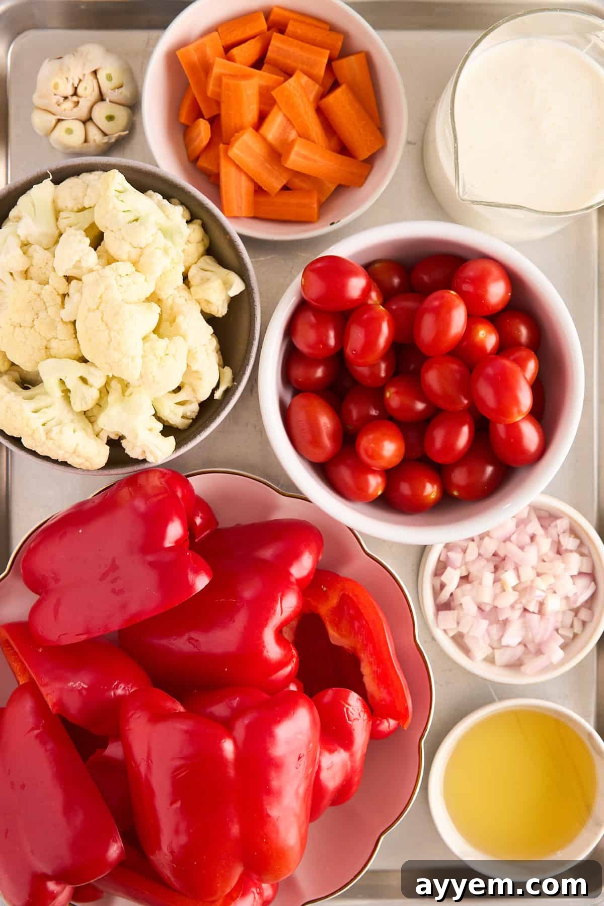 A vibrant selection of fresh, uncooked ingredients displayed neatly on a white tray, including bright orange carrots, creamy white cauliflower florets, plump red grape tomatoes, whole red bell peppers, diced shallots, and a whole garlic bulb, ready for roasting. A small bowl of olive oil and a glass of creamy liquid (coconut milk) are also present.