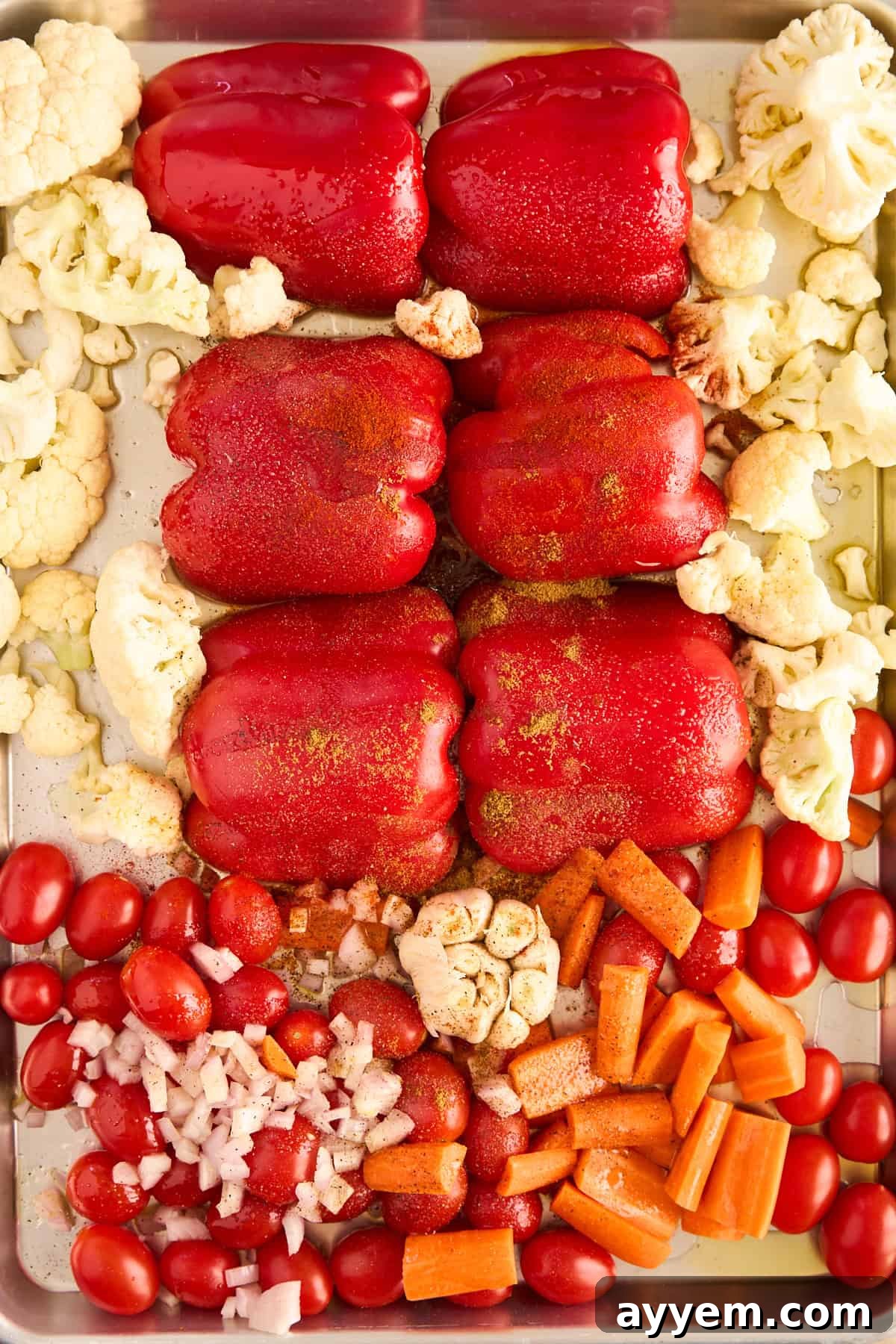 A baking tray filled with fresh, uncooked red bell peppers, cauliflower florets, grape tomatoes, chopped onions, carrot pieces, and whole garlic, all seasoned with herbs and oil and arranged in neat sections, ready for roasting to create a flavorful soup.
