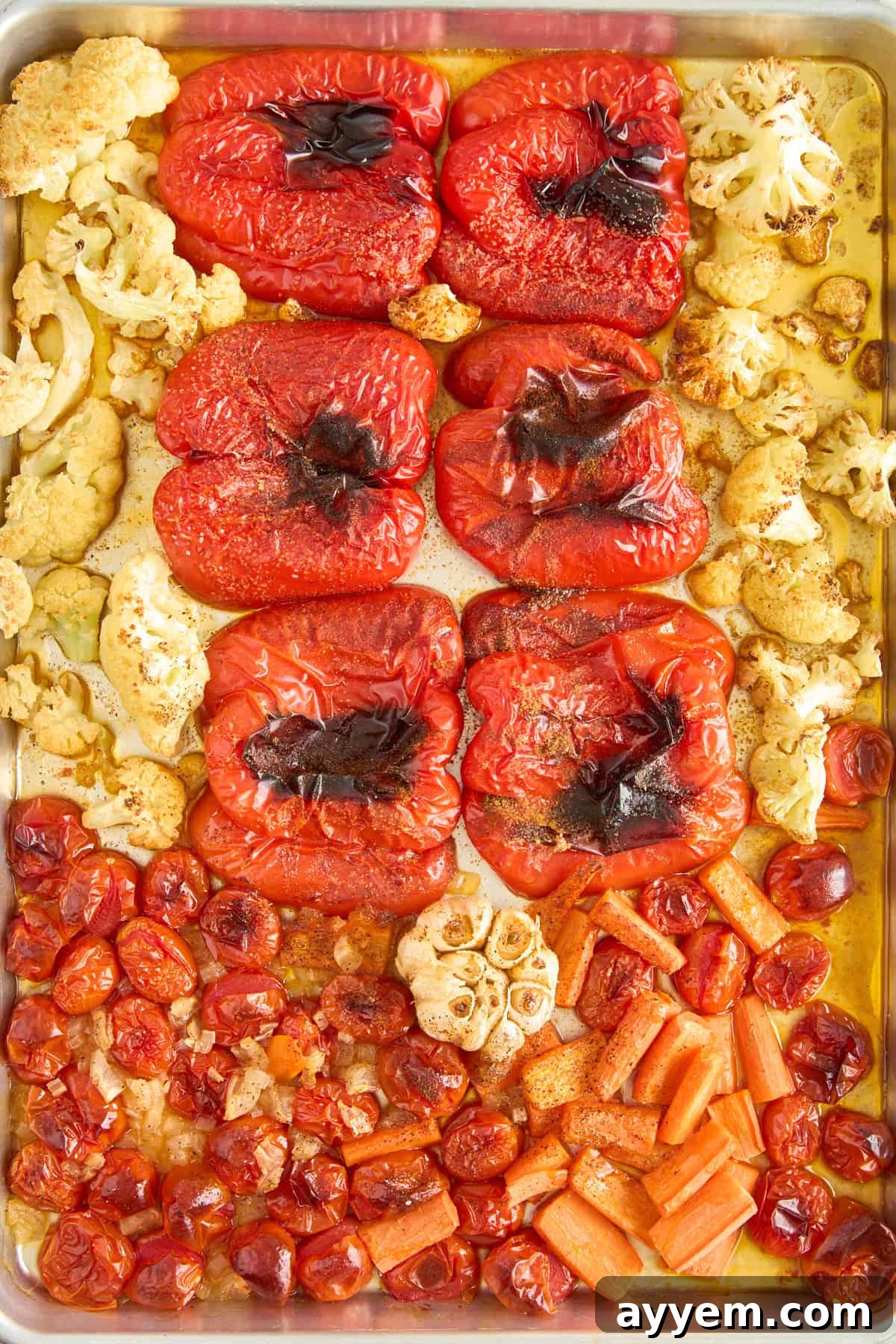 A close-up shot of a baking sheet featuring roasted red bell peppers with slightly charred skins, golden-brown cauliflower florets, blistered cherry tomatoes, tender whole garlic cloves, and softened chopped carrots, all glistening with oil after being cooked in the oven.
