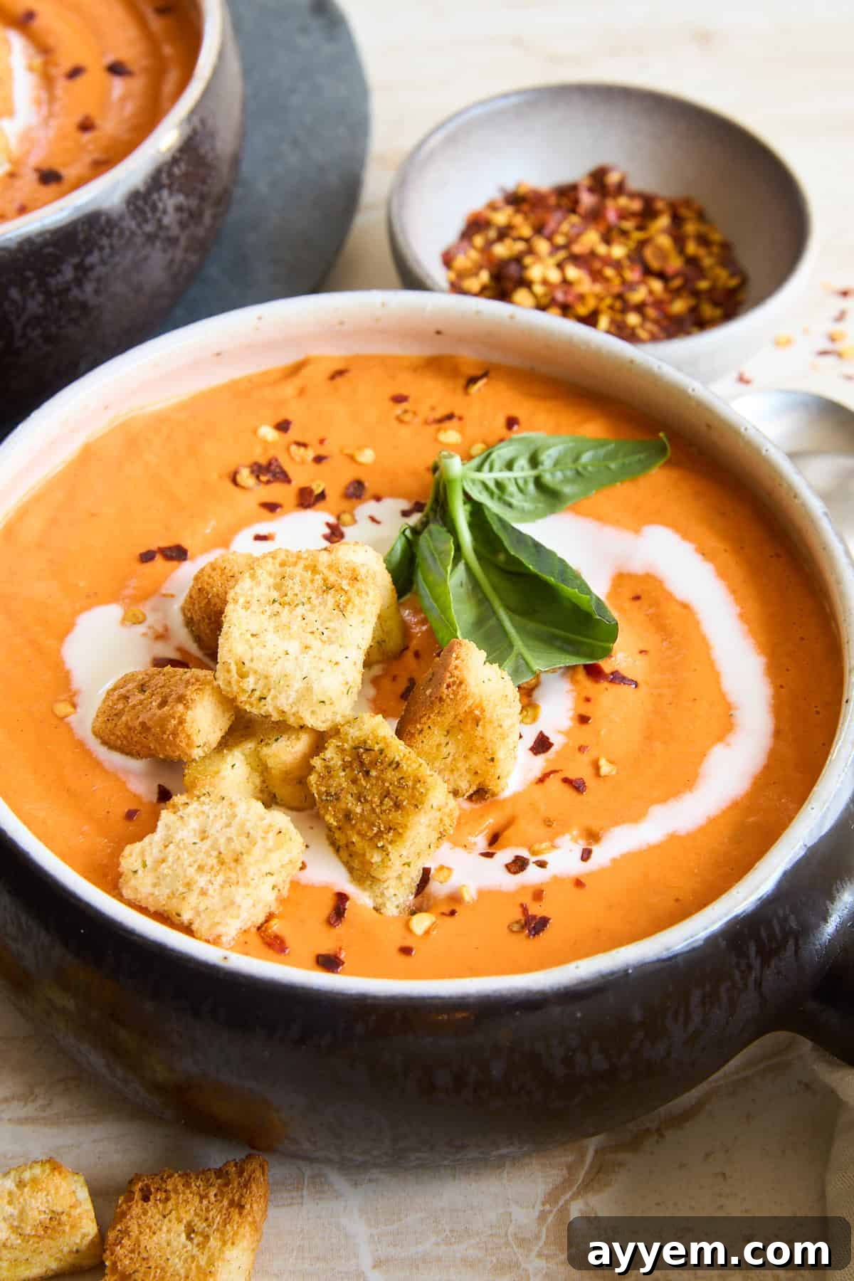 A beautifully presented bowl of creamy roasted red pepper and cauliflower soup, garnished with crispy croutons, a vibrant swirl of coconut cream, and fresh basil leaves. A small dish of red pepper flakes sits in the background.