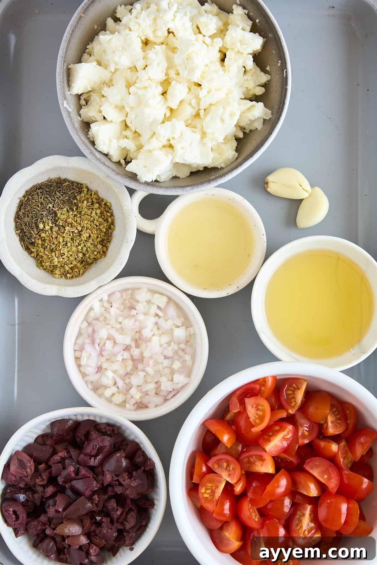 A kitchen counter showcasing a variety of fresh ingredients prepped for cooking, including bowls of crumbled feta cheese, halved cherry tomatoes, finely chopped shallots (though not explicitly in this recipe, it's often a Mediterranean staple), sliced black olives, dried oregano, a bottle of olive oil, a glass of white wine, and two cloves of garlic, suggesting a flavorful and aromatic meal in progress.