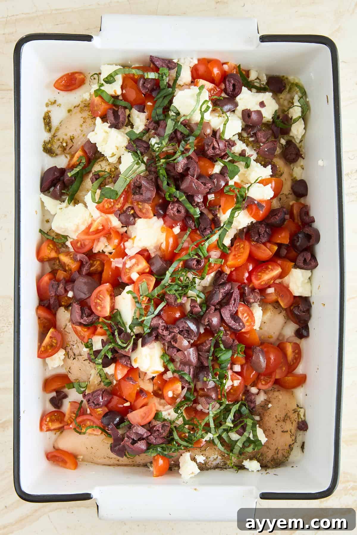 A pristine baking dish showcasing raw chicken breasts meticulously arranged and topped with halved cherry tomatoes, generous crumbles of feta cheese, an array of sliced black olives, and delicate strips of fresh basil, all ready for the oven to transform them into a delicious meal.