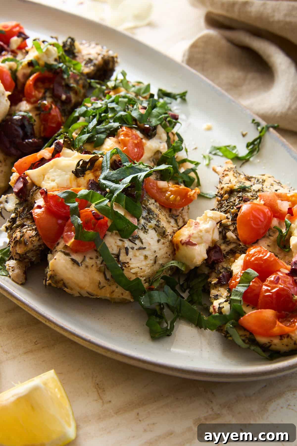 A beautifully plated herb-seasoned baked chicken breast, generously topped with a vibrant mixture of chopped tomatoes, briny olives, crumbled feta cheese, and fresh basil leaves, served on an elegant white oval plate. A bright lemon wedge is visible in the corner, adding a fresh, zesty touch to the Mediterranean-inspired meal.