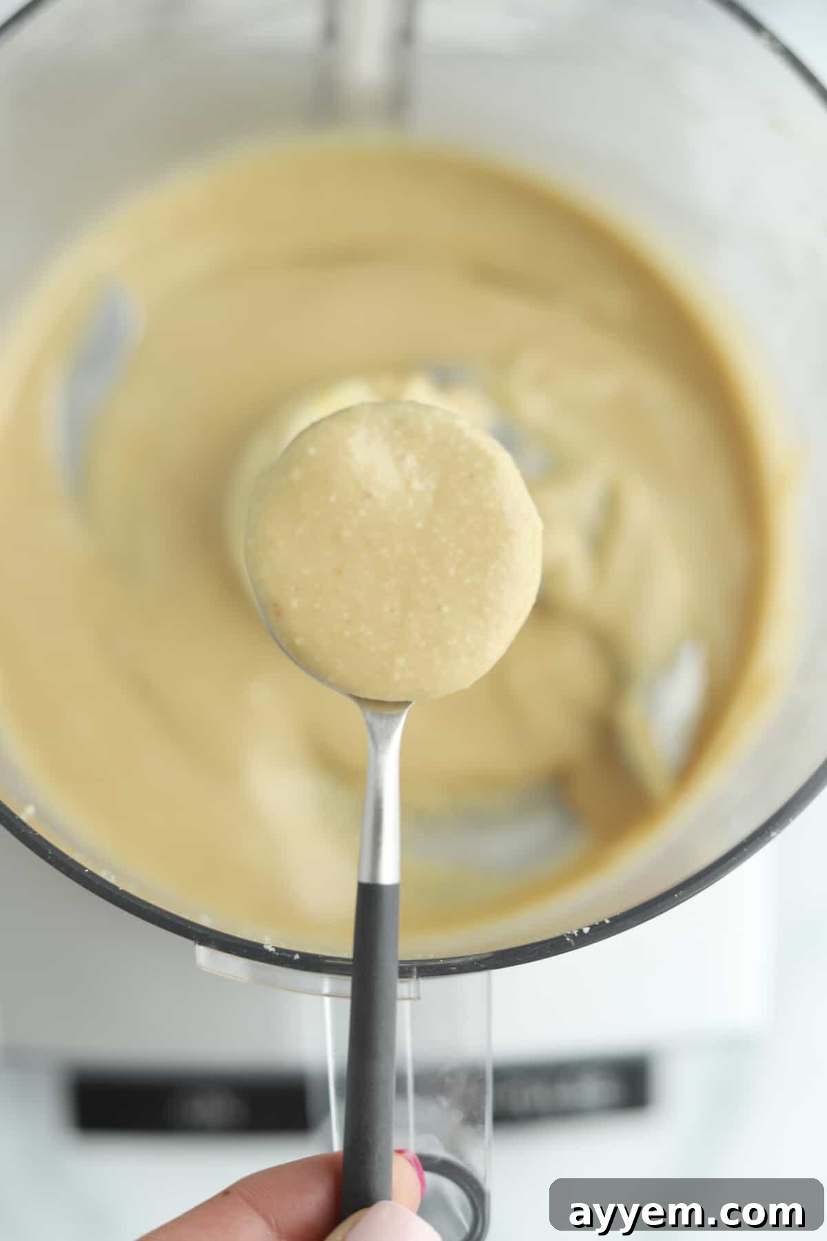 A spoon lifting a dollop of smooth hazelnut butter over a food processor bowl filled with more hazelnut butter.