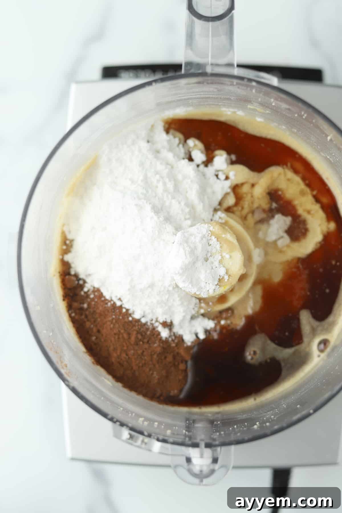 Melted chocolate chips, brewed espresso, powdered sugar, vanilla extract, cocoa powder, and water, artfully arranged around a food processor containing hazelnut butter, showcasing the ingredients ready for blending.