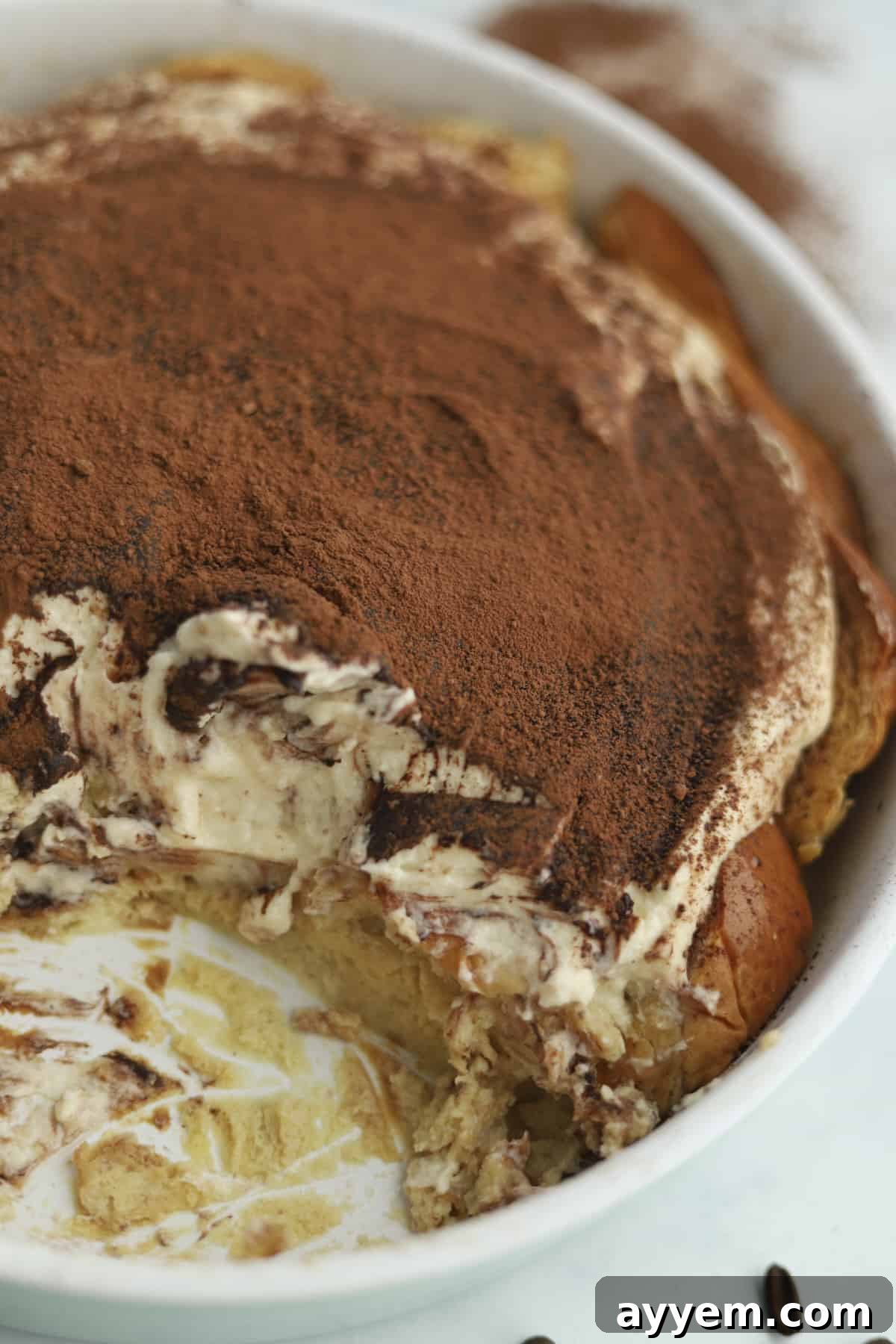 A large portion of Tiramisu Bread Pudding has been removed from a baking dish, revealing its creamy, layered interior, topped with a dusting of cocoa powder.