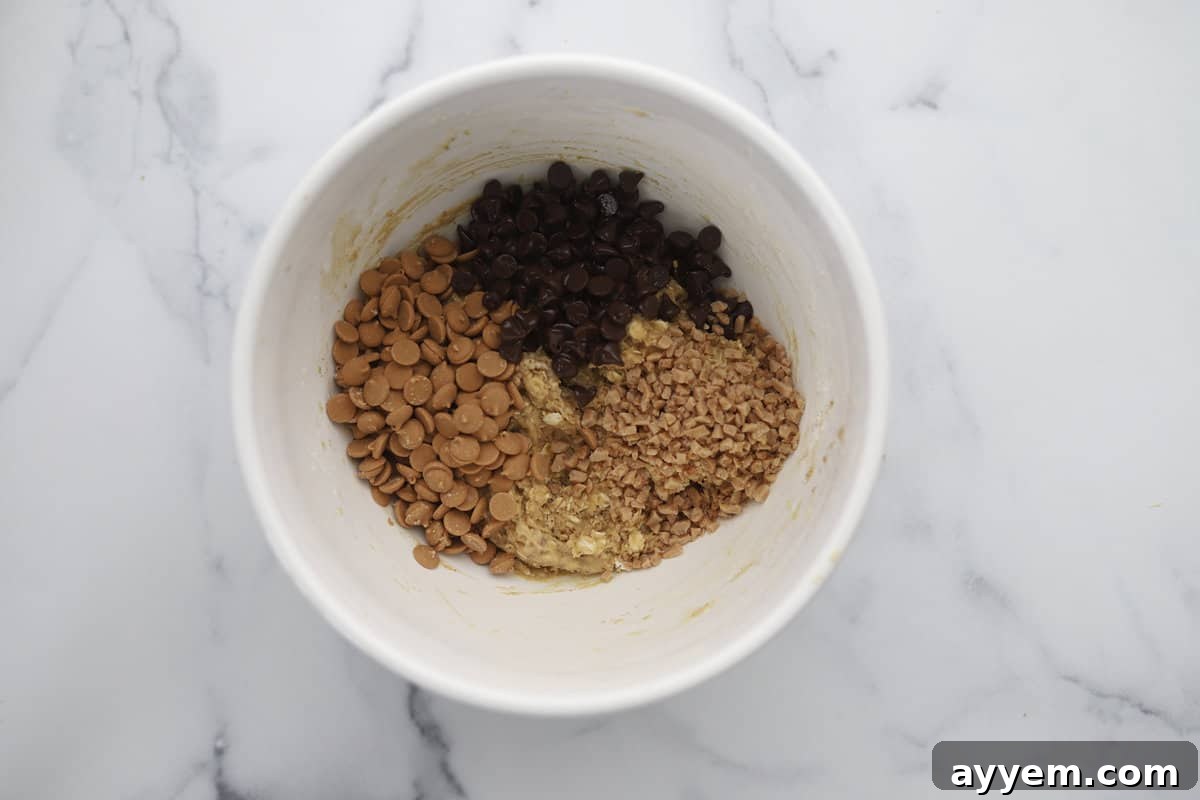 A close-up of chocolate chips, peanut butter chips, and toffee pieces being carefully folded into the rich cookie dough batter.