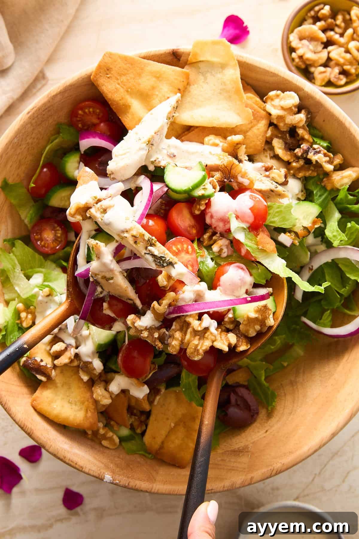 A person using wooden salad tongs to serve a fresh Whipped Feta Chicken Salad from a large wooden bowl, showcasing crisp lettuce, colorful cherry tomatoes, cucumber, red onion, walnuts, and pita chips, all generously coated with creamy dressing. A small bowl of walnuts is placed nearby.