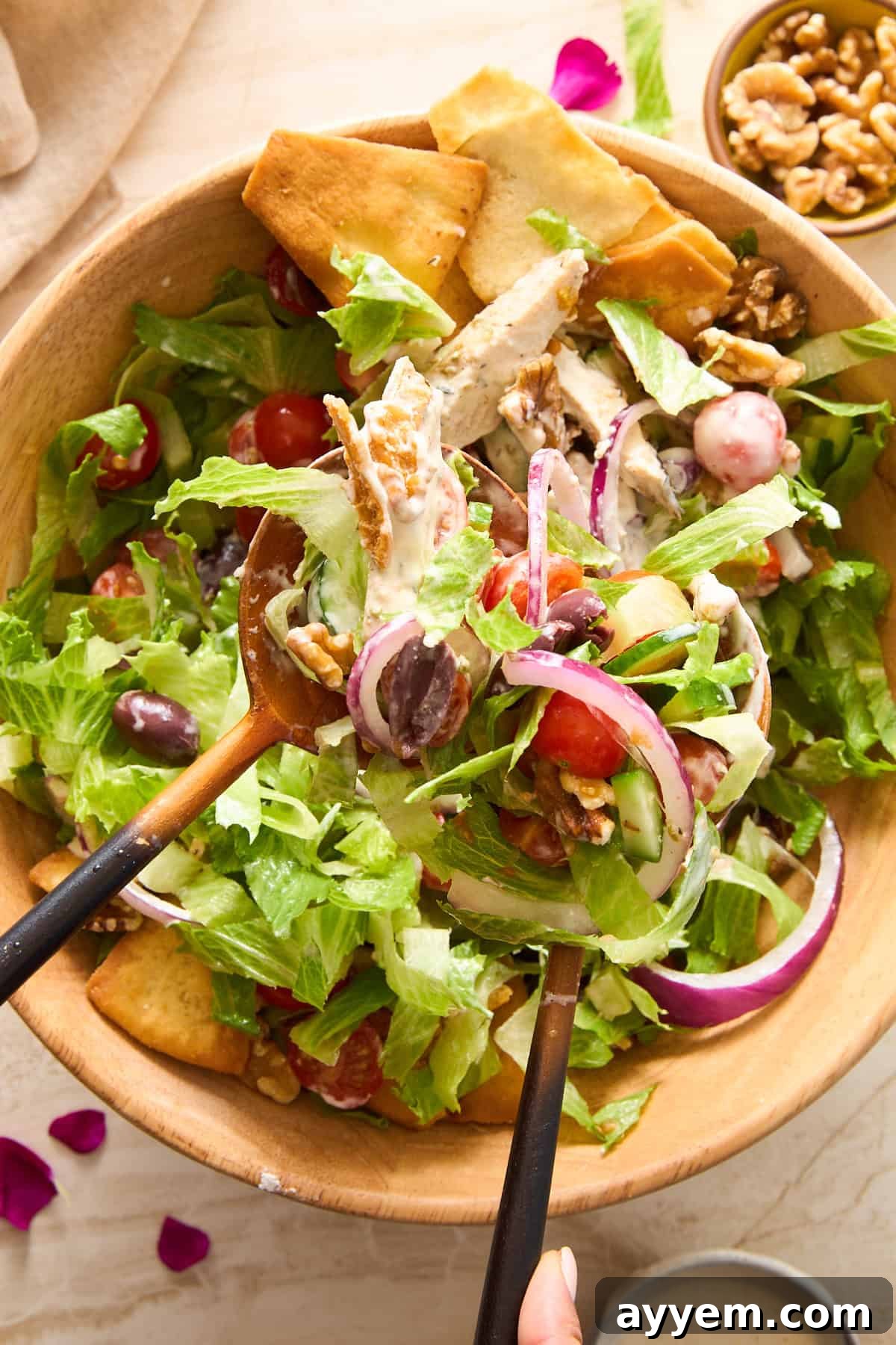 Two wooden serving spoons are tossing a vibrant Whipped Feta Chicken Salad in a large wooden bowl. The salad includes crisp lettuce, juicy grape tomatoes, thinly sliced red onion, briny olives, crunchy walnuts, and golden pita chips, all generously coated in a creamy dressing. A small bowl of walnuts is visible in the background.