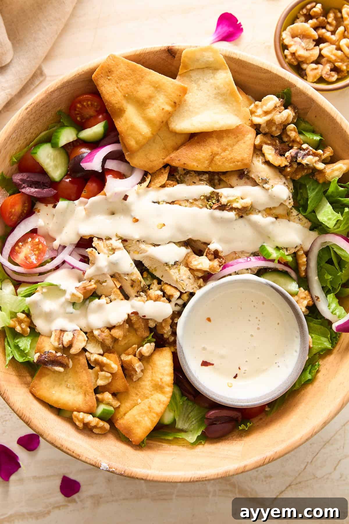A wooden bowl filled with salad greens, cherry tomatoes, cucumber, red onion, pita chips, walnuts, olives, and drizzled with creamy dressing. A small bowl of extra dressing sits inside the salad.