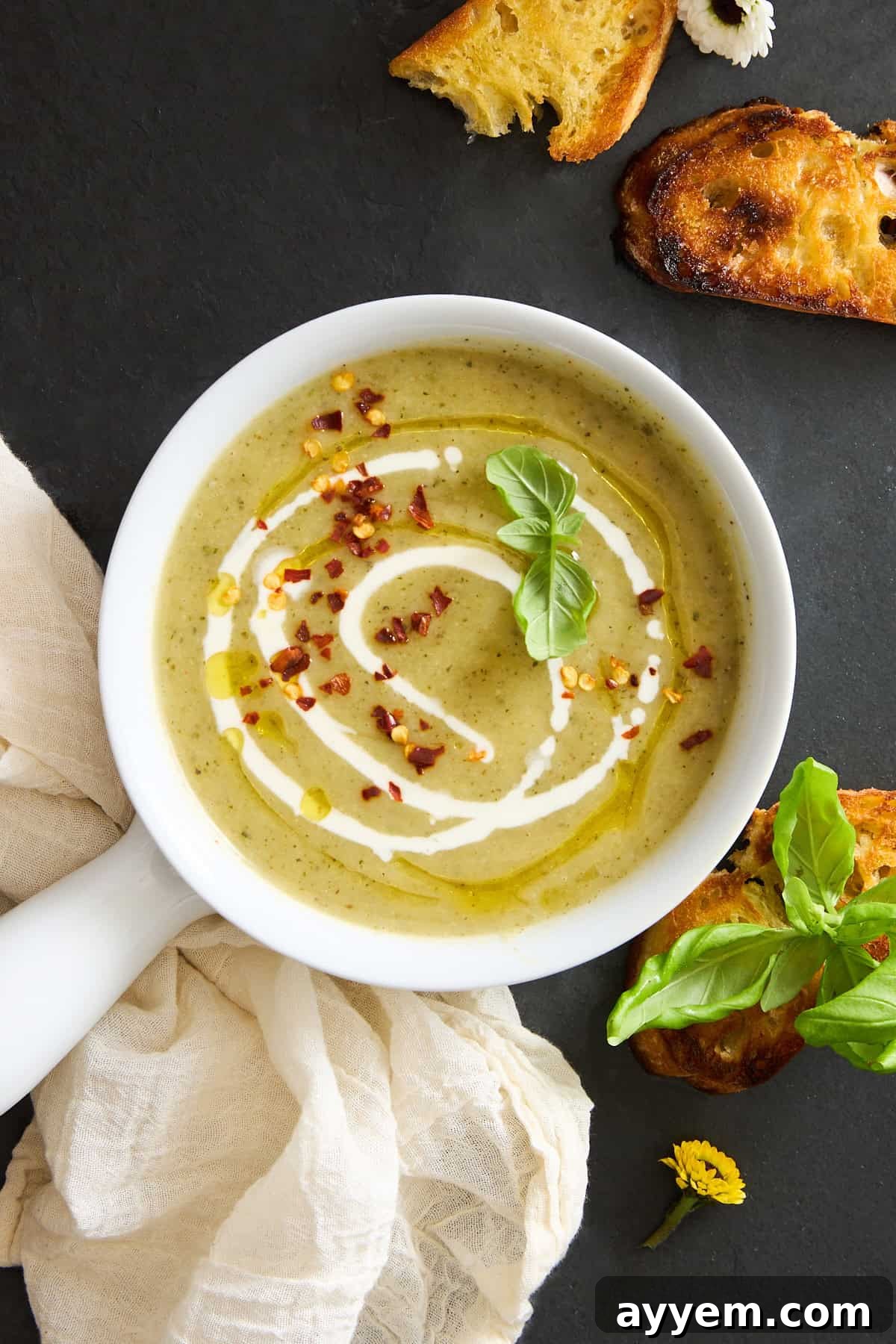 A beautifully presented bowl of vibrant green creamy zucchini soup, artfully garnished with a swirl of fresh cream, a sprinkle of fiery red chili flakes, a glistening drizzle of high-quality olive oil, and fragrant fresh basil leaves. It is served alongside perfectly toasted bread slices, arranged on a rustic dark surface, with a soft, light-colored cloth and a cheerful yellow flower adding to the inviting scene.