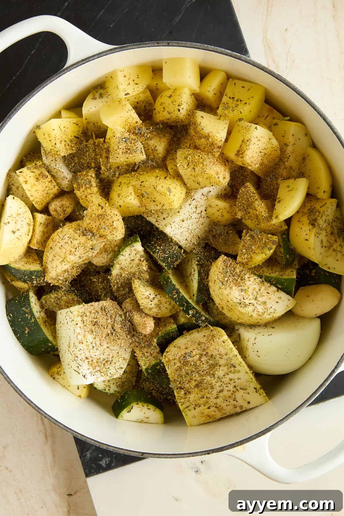 A sturdy white pot brimming with a colorful assortment of freshly chopped vegetables, including tender potatoes, vibrant zucchini, pungent onions, and whole garlic cloves. All are generously seasoned with a medley of dried herbs and freshly ground black pepper, sitting ready for cooking on a pristine marble countertop.