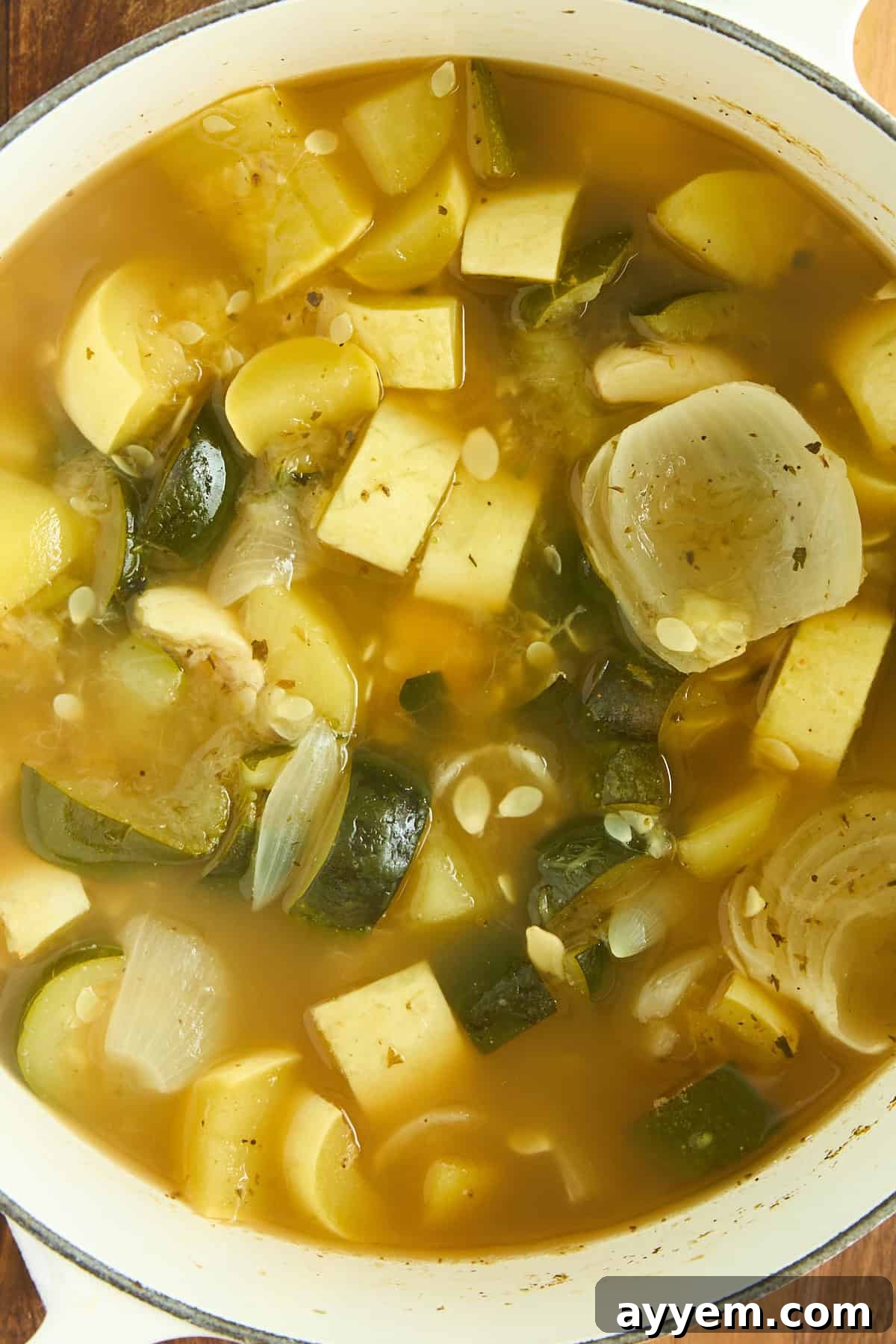 A close-up view of a pot filled with simmering vegetable soup, featuring tender chunks of onion, vibrant zucchini, and other aromatic vegetables suspended in a clear, flavorful broth, ready for blending.