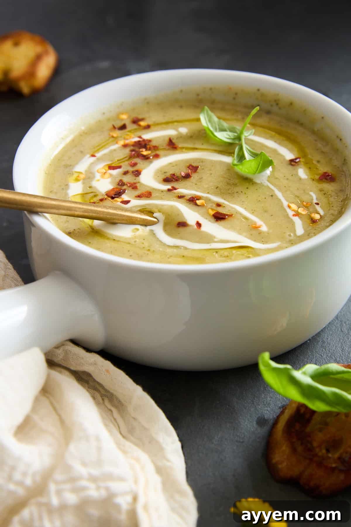 A delectable creamy green soup beautifully presented in a white bowl, exquisitely garnished with a delicate swirl of fresh cream, a sprinkle of vivid red pepper flakes, and a verdant basil leaf, all finished with a shimmering drizzle of olive oil. A elegant gold spoon rests gently inside the bowl, which sits on a dark, rustic surface beside a soft, light cloth and slices of golden toasted bread, creating an inviting and appetizing tableau.