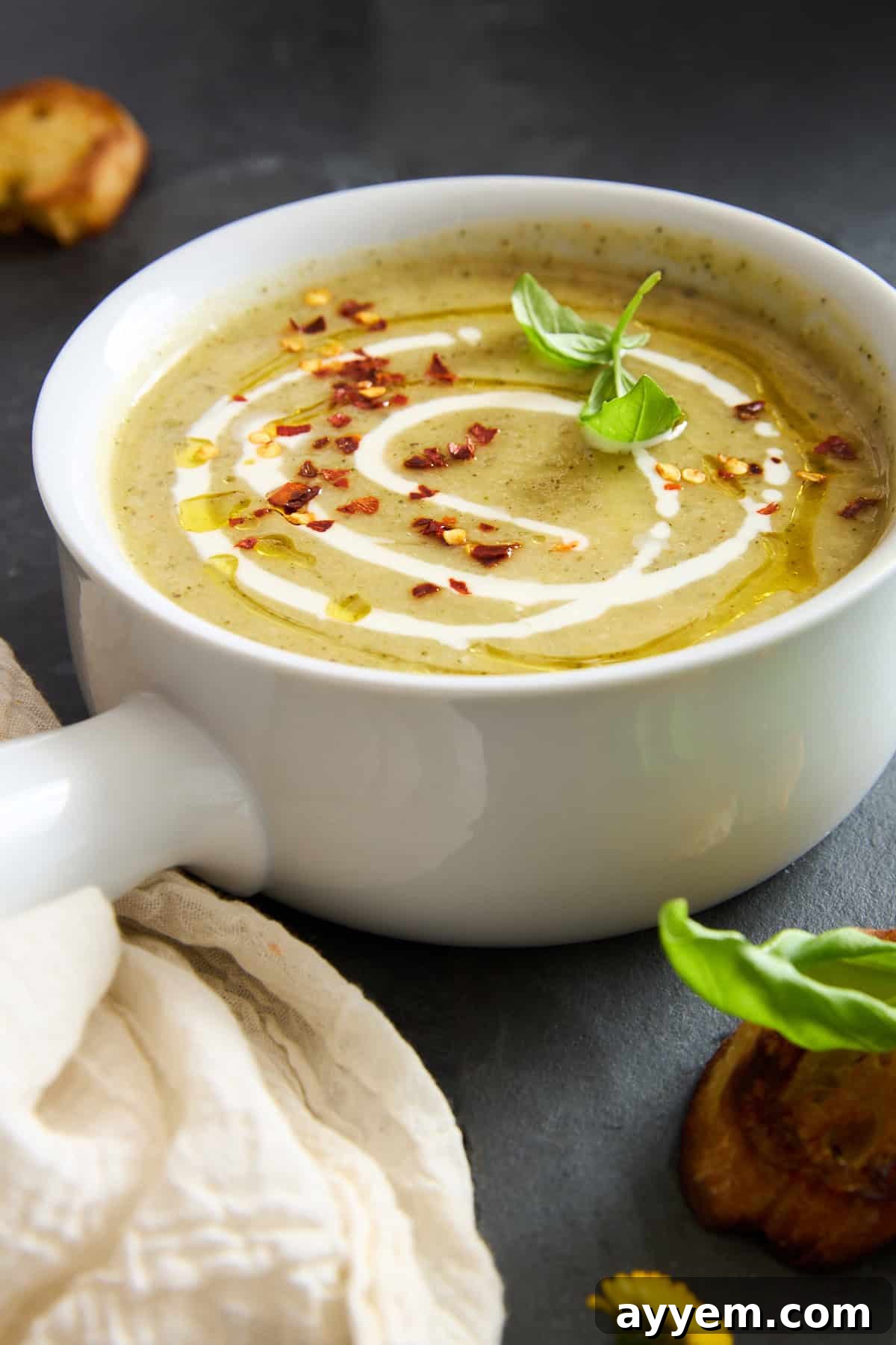 A bowl of rich creamy zucchini soup, garnished with a swirl of cream, red pepper flakes, a basil leaf, and a drizzle of oil, served in a pristine white bowl with a handle on a dark, rustic surface.