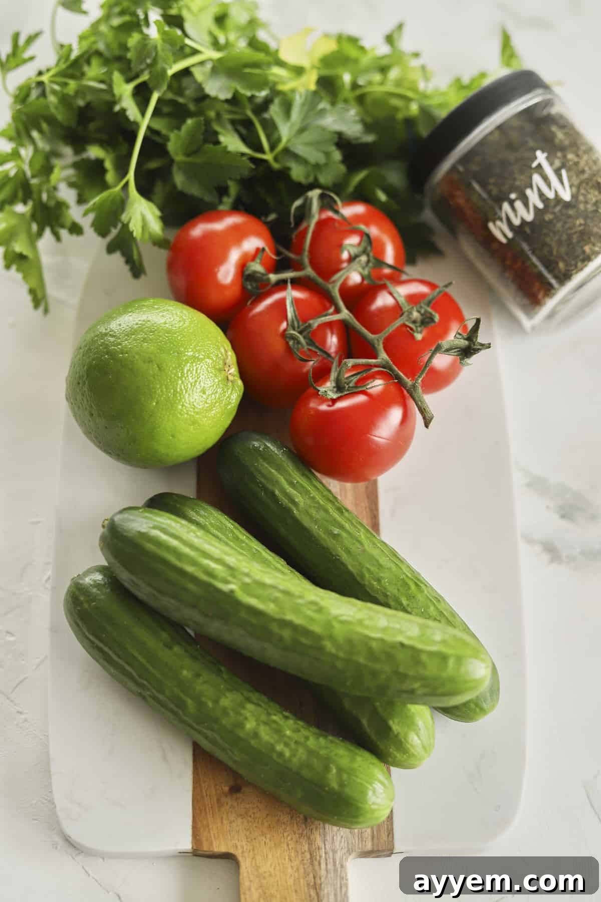Ingredients for Egyptian Tomato & Cucumber Salad: whole cucumbers, tomatoes on the vine, a lime, fresh cilantro, and fresh mint sprigs arranged on a rustic surface.