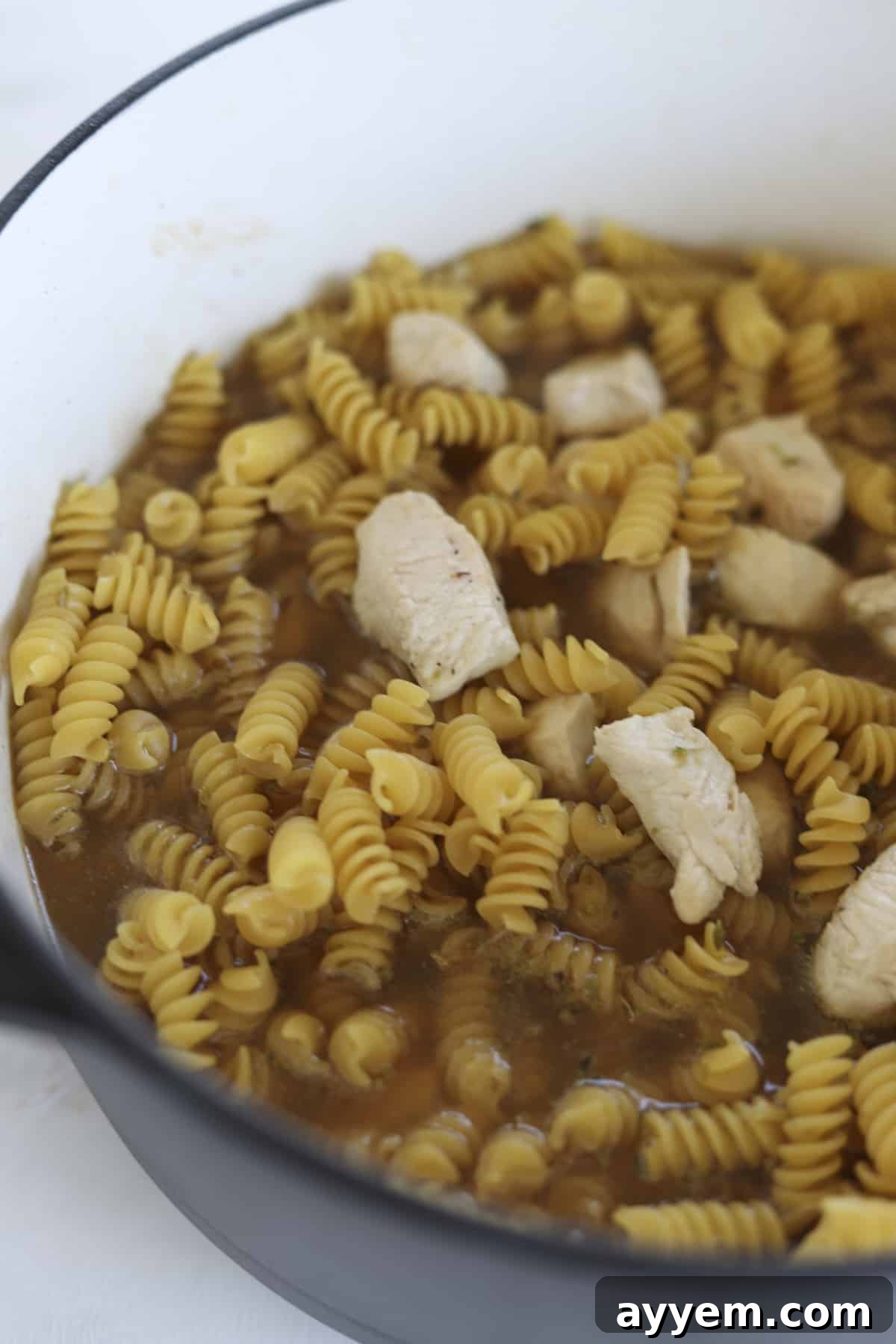 Uncooked rotini pasta and chicken simmering in savory broth within a single pot, absorbing flavors.