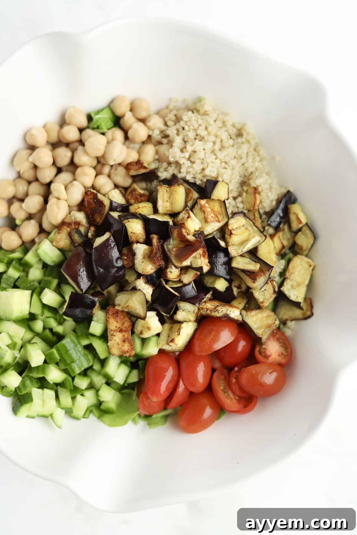Overhead view of quinoa, chickpeas, roasted eggplant, cucumber, and tomatoes in a bowl ready to be combined for roasted eggplant salad