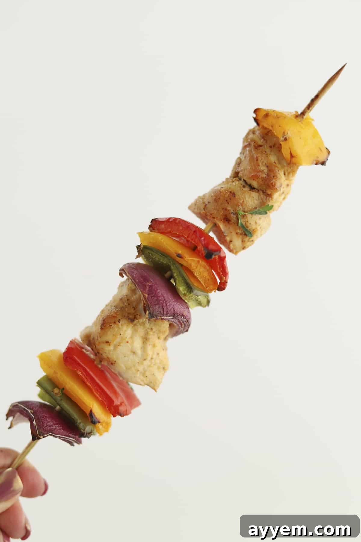 a hand holding an oven-baked chipotle chicken skewer with red, yellow, and green peppers and red onion