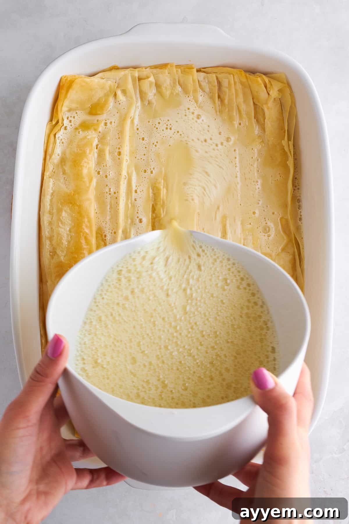 Egg and milk mixture being poured over partially baked crinkled phyllo dough in a baking dish.