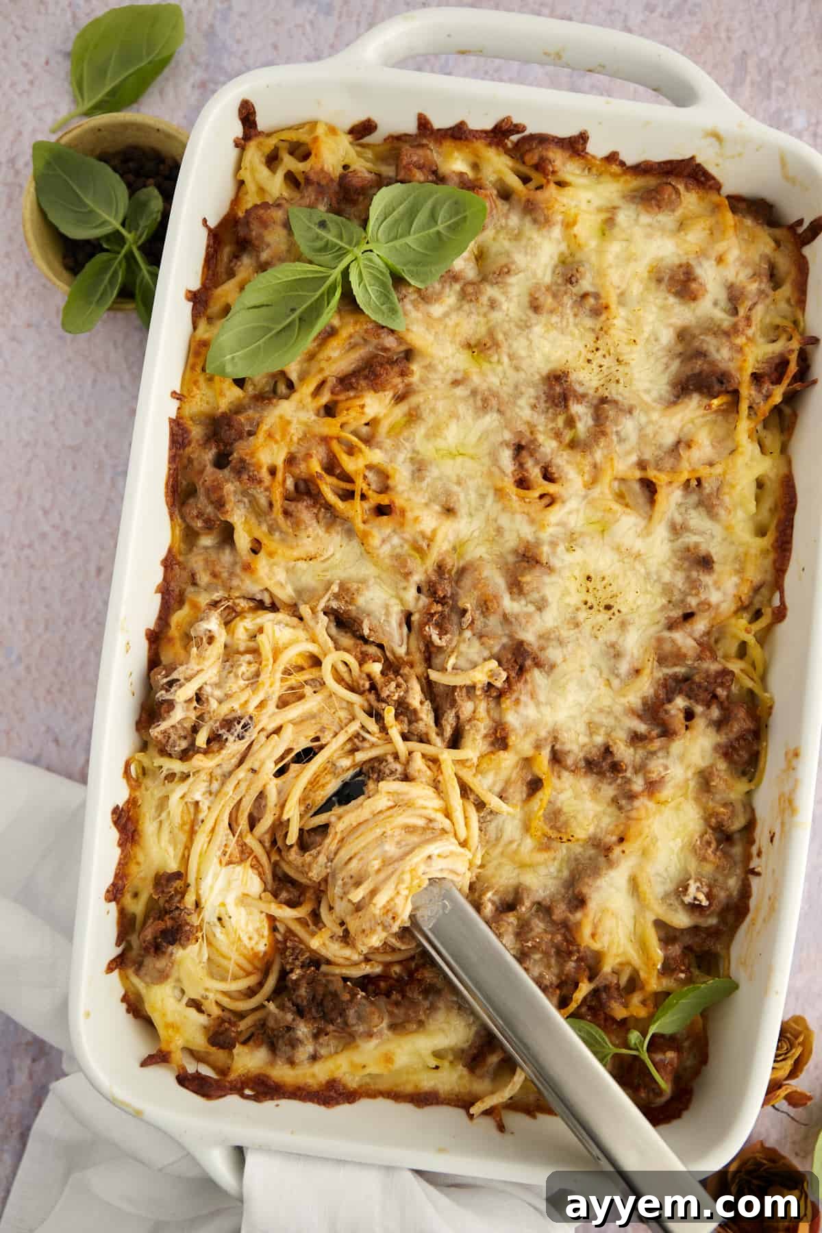 Tongs gracefully twirling a perfect serving of baked Million Dollar Spaghetti casserole, showcasing its rich layers and golden cheese topping, in a baking dish.
