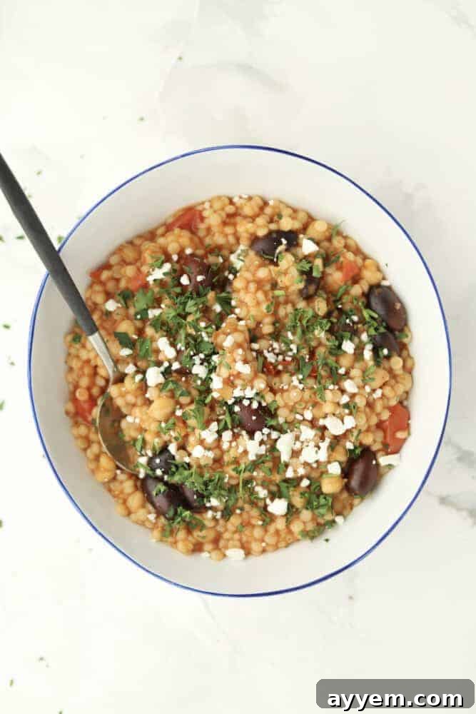 a bowl of Greek couscous topped with crumbled feta and fresh herbs with a spoon sticking out 