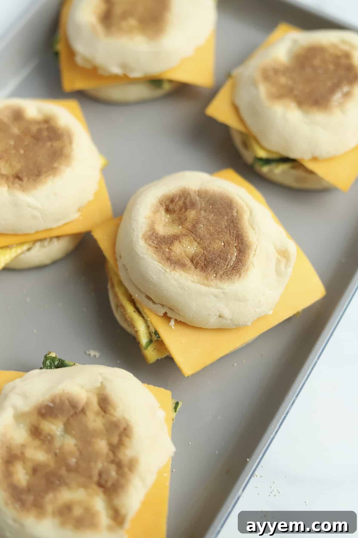 Arranged protein-packed egg muffin sandwiches topped with raw American cheese slices on a baking dish, perfectly aligned and ready for the oven to melt the cheese.