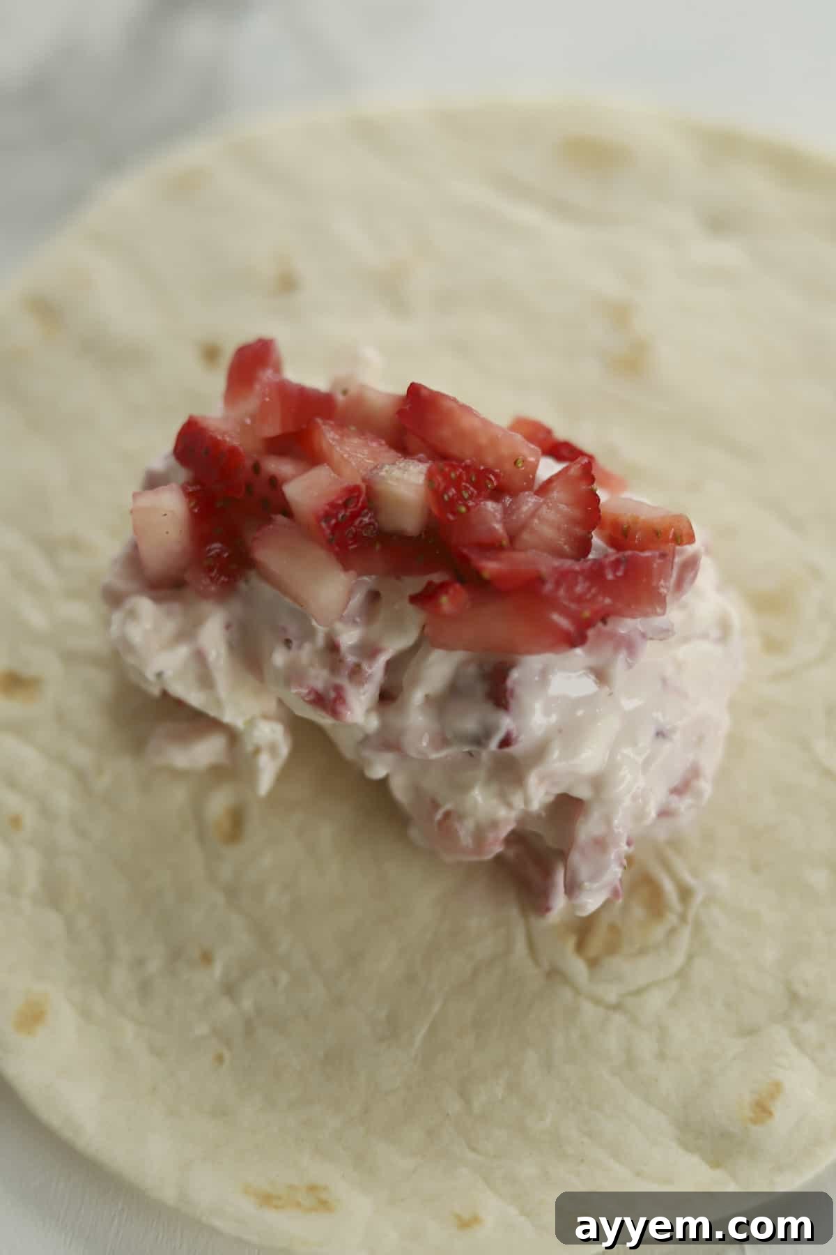 A soft flour tortilla shell with a generous scoop of strawberry cream cheese mixture carefully placed in the center, ready for folding.