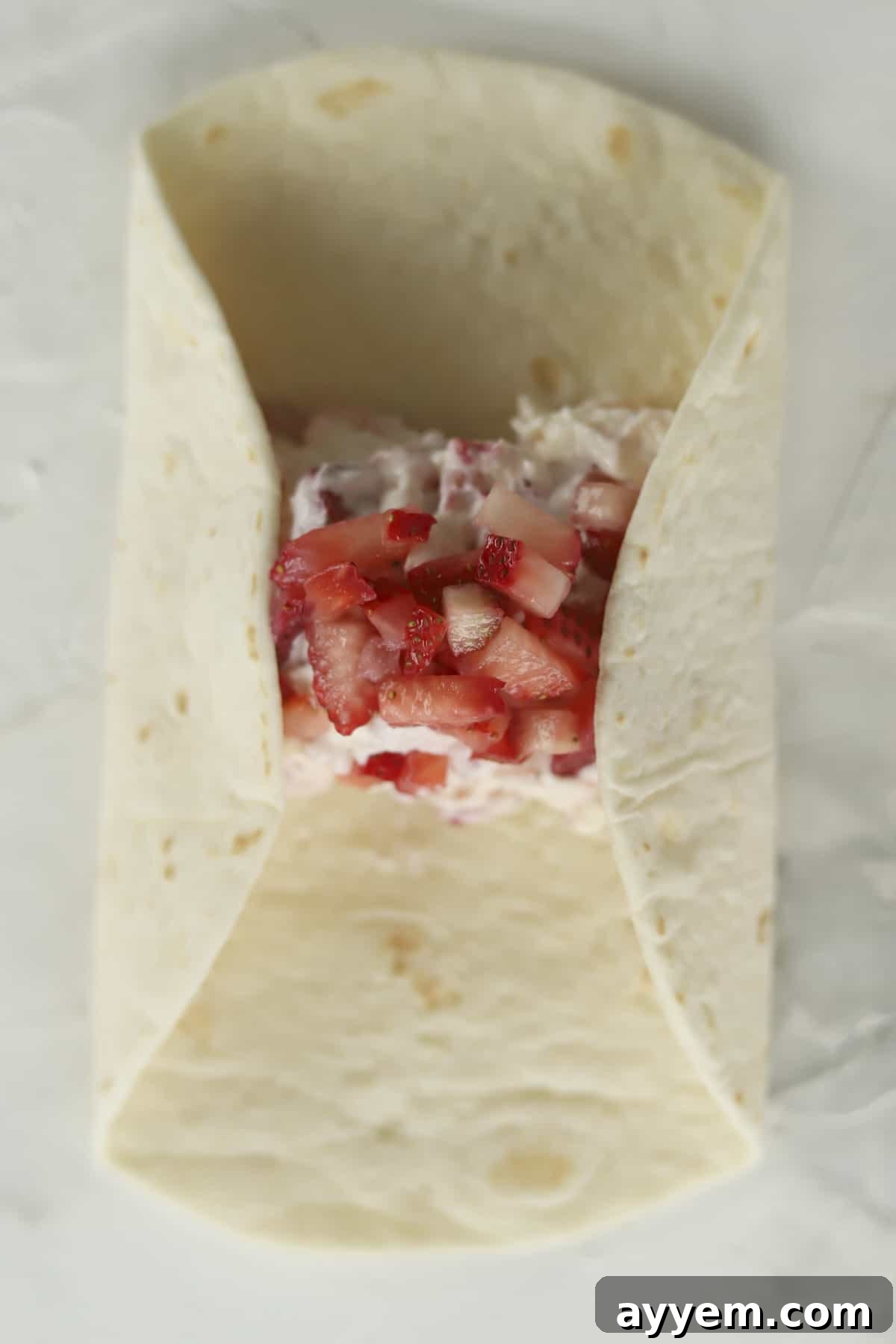 Hands demonstrating the folding technique of a tortilla, carefully tucking in the sides over the strawberry cheesecake filling to create a secure burrito shape.