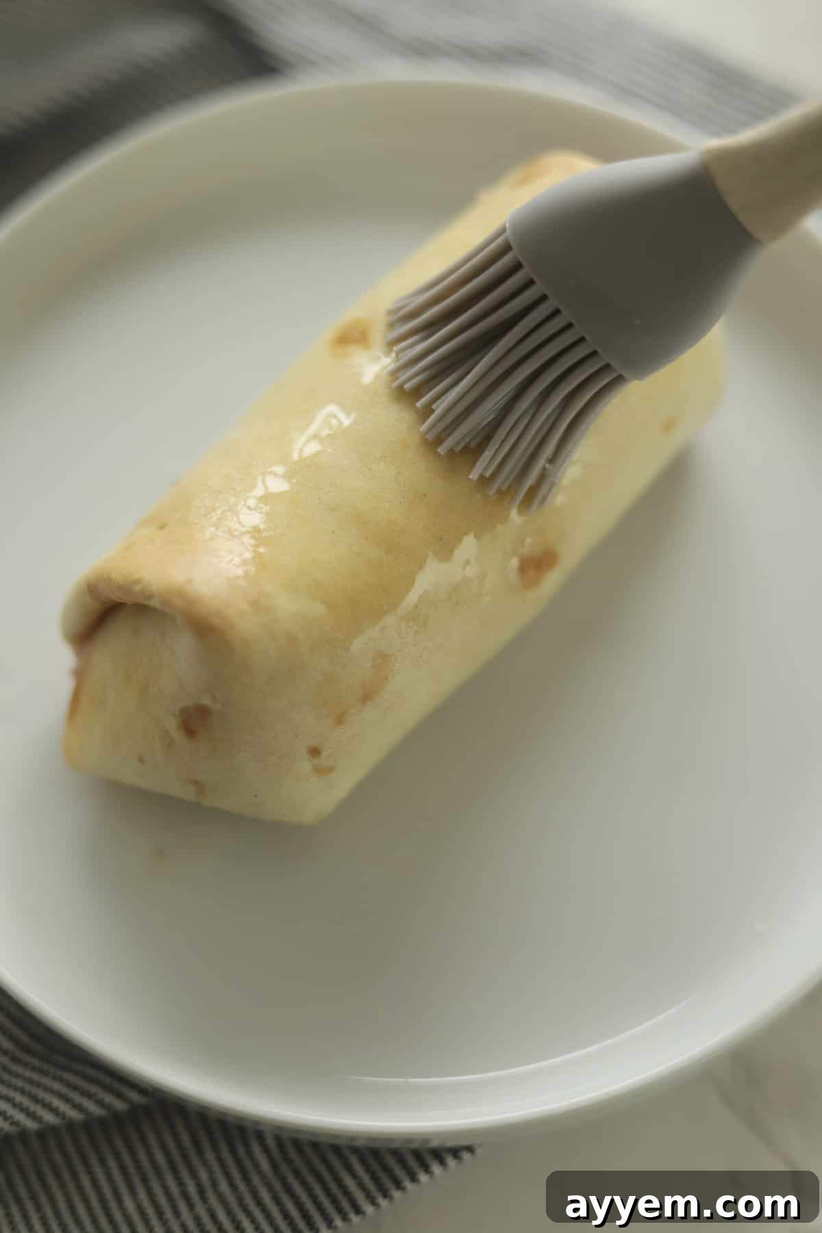 A neatly rolled strawberry cheesecake chimichanga being brushed with melted butter before its final dusting of cinnamon sugar.
