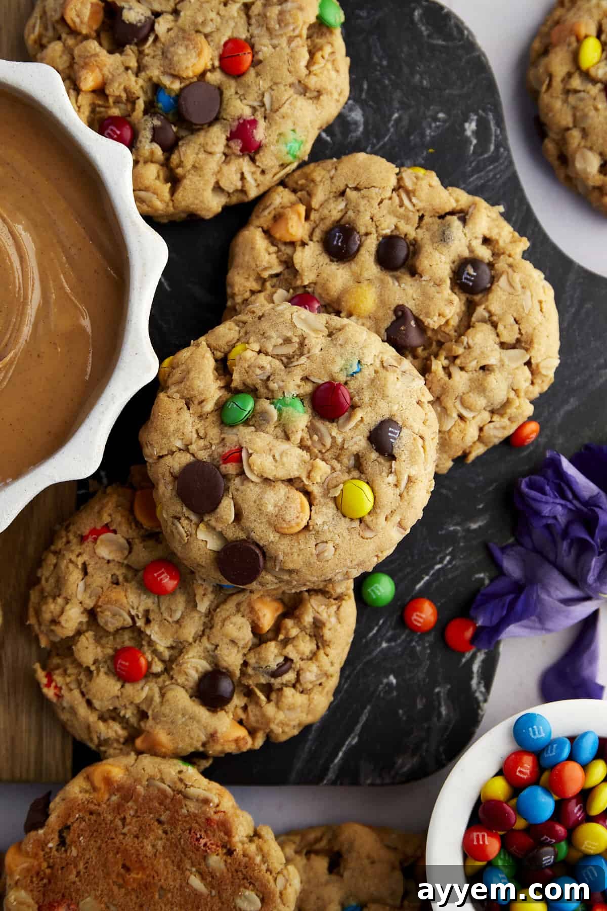 Fully Loaded Monster Cookies 2 A vibrant display of freshly baked Monster Cookies next to bowls brimming with creamy peanut butter and colorful M&M's, ready to be enjoyed.