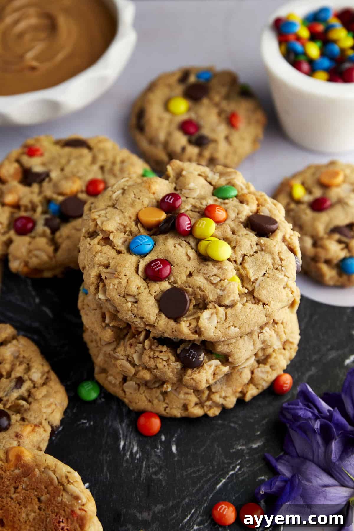 Fully Loaded Monster Cookies 4 An array of delicious Monster Cookies adorned with M&M's and chocolate chips, with the vibrant ingredients like peanut butter and M&M's artfully blurred in the background.