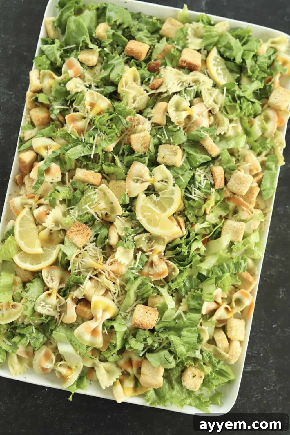 overhead image of a white serving platter full of pesto pasta salad made with lettuce, noodles, croutons, and Parmesan