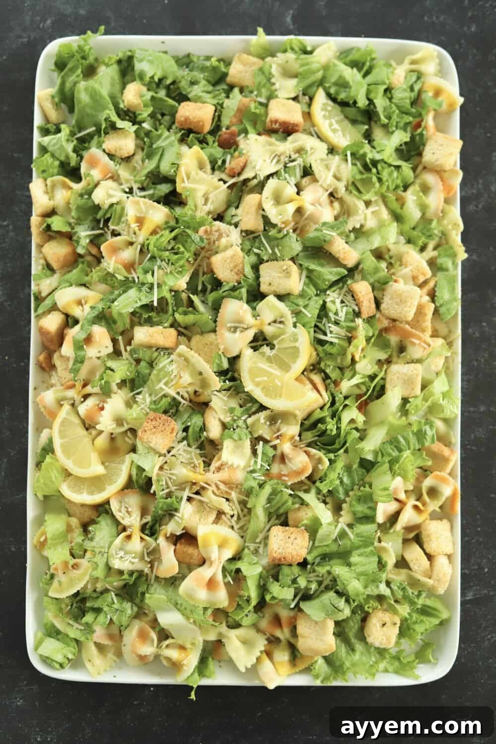 a rectangular white serving platter full with pesto pasta salad made with lettuce, noodles, Parmesan, croutons, and a pesto sauce topped with lemon wedges