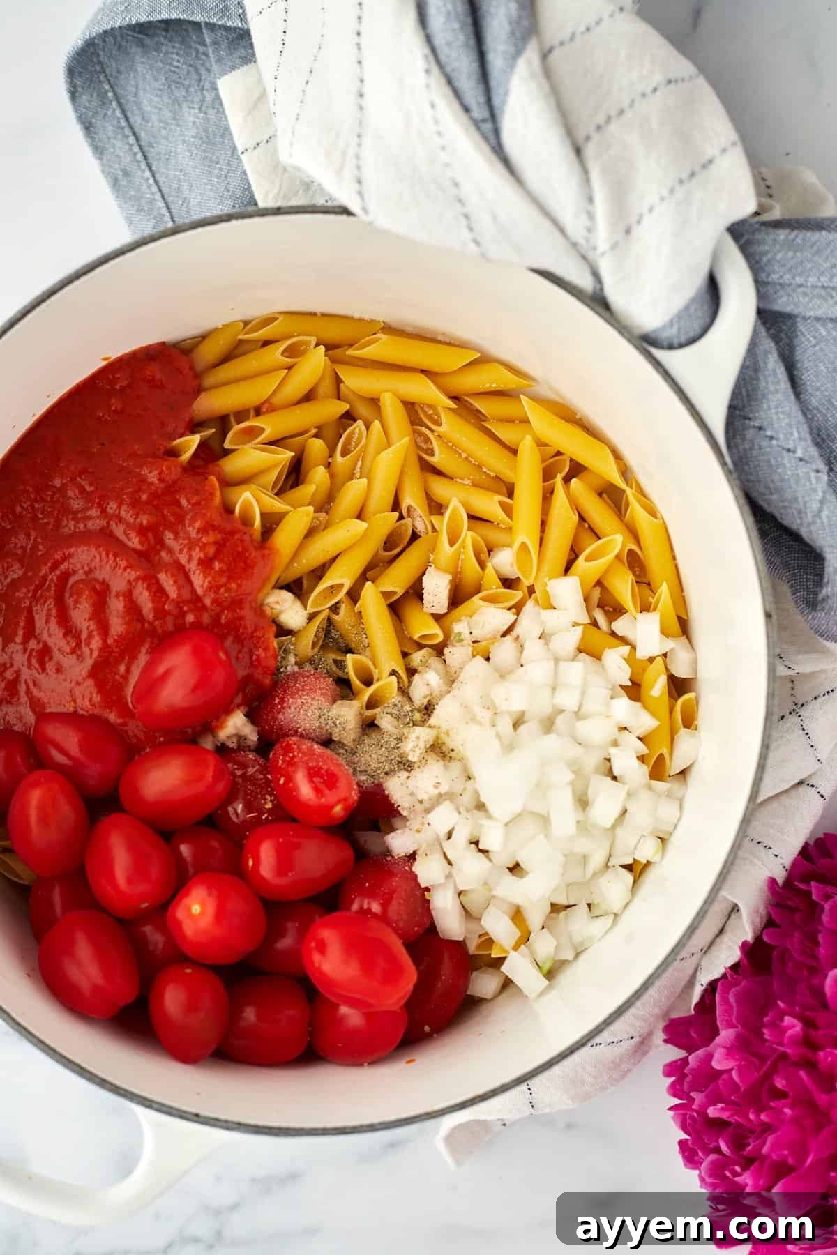 A large pot filled with uncooked penne pasta, vibrant grape tomatoes, chopped onions, marinara sauce, and fresh herbs, ready for cooking.