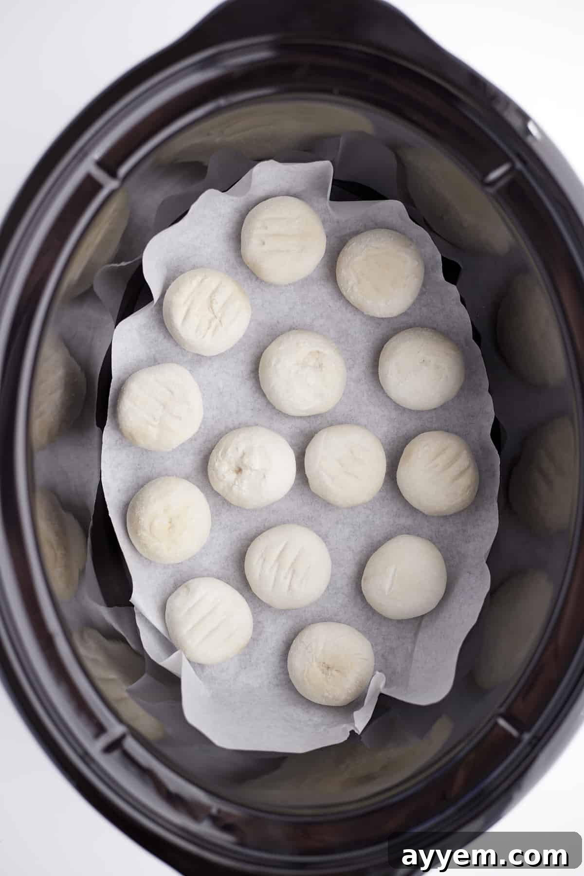 frozen dinner rolls arranged in the bottom of a parchment lined slow cooker, showing the raw 'secret ingredient'