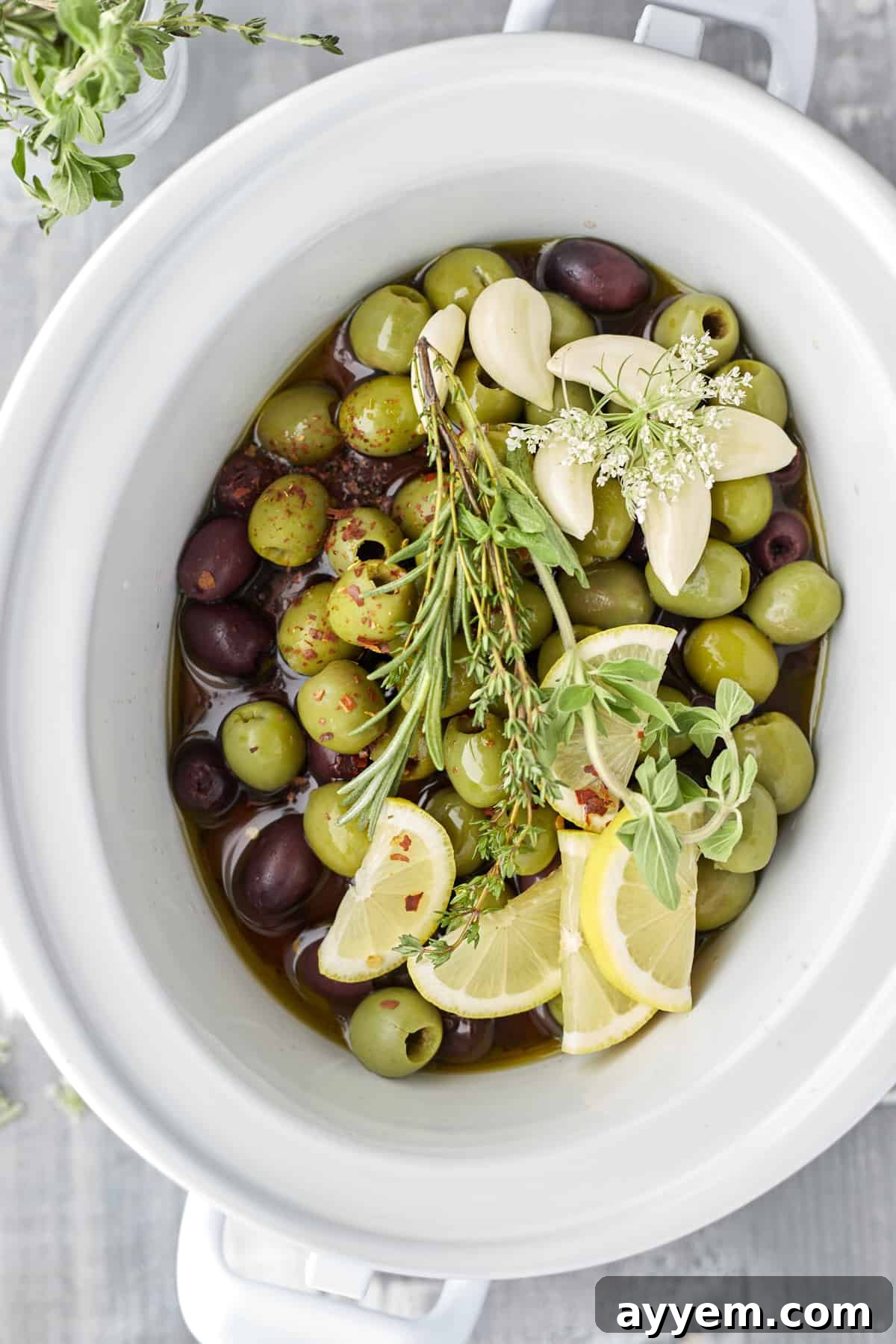 overhead image of green and red olives, lemon wedges, garlic, and herbs in a slow cooker to make roasted olives