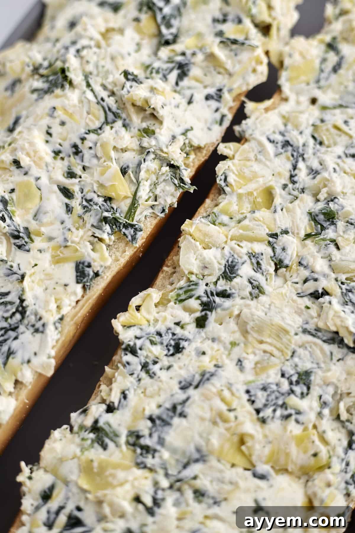 Loaded Garlic Bread with Spinach and Artichokes 4 Two halves of French bread generously topped with a creamy spinach and artichoke mixture, ready for baking to become homemade garlic bread.