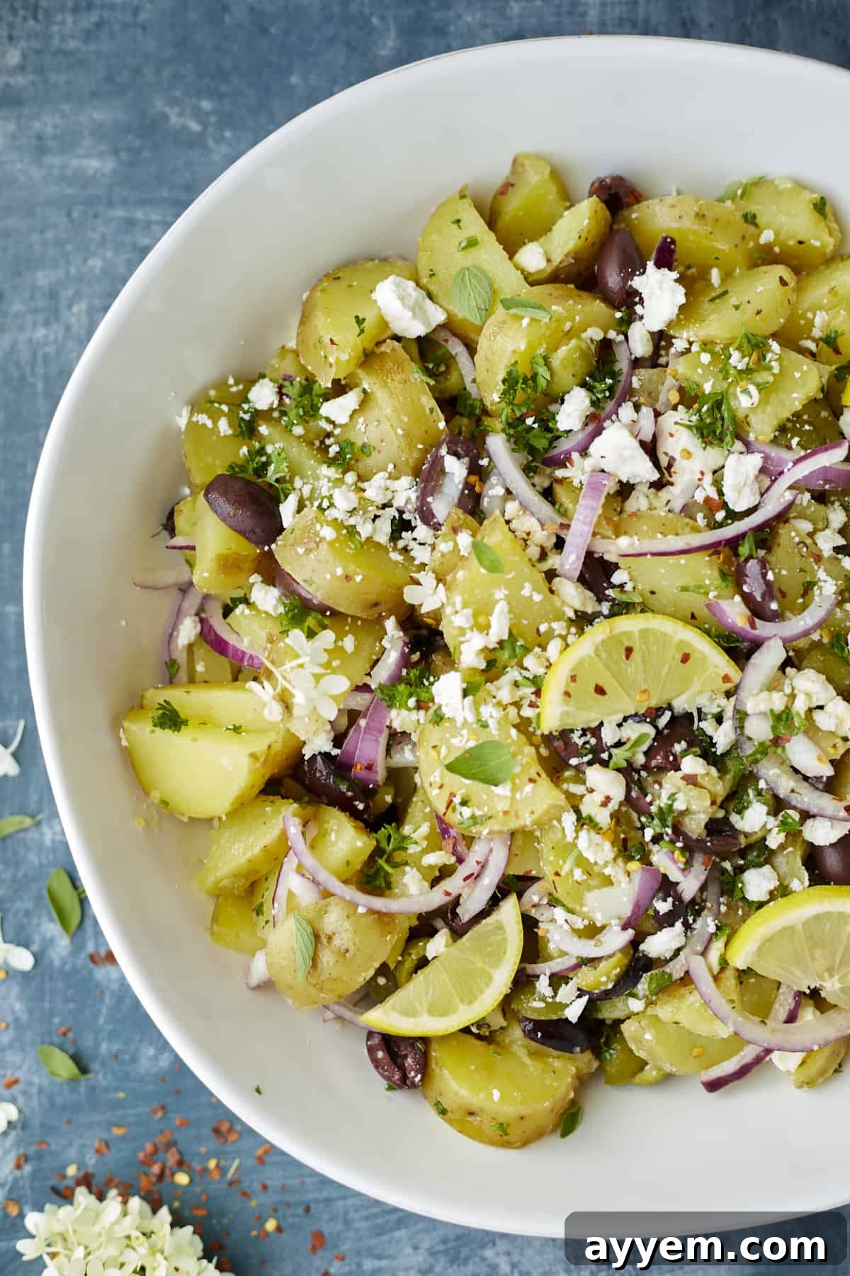 A beautifully presented bowl of Lemon Greek Potatoes, adorned with fresh red onions, assorted olives, crumbled feta, fragrant fresh herbs, and bright lemon wedges, ready to serve