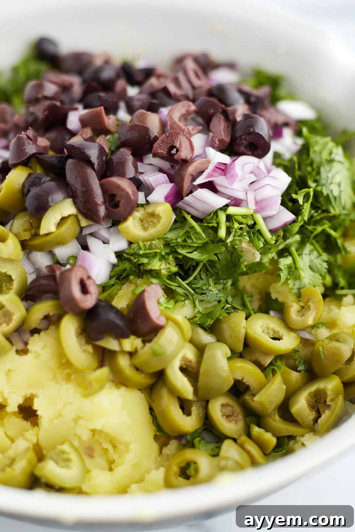 overhead image of yukon gold mashed potatoes topped with garlic, olives, and herbs ready to be stirred.