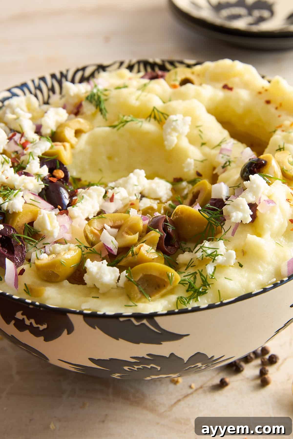 A bowl of mashed potatoes topped with chopped green and black olives, crumbled feta cheese, red onion, dill, and red pepper flakes, in a decorative black and white bowl.