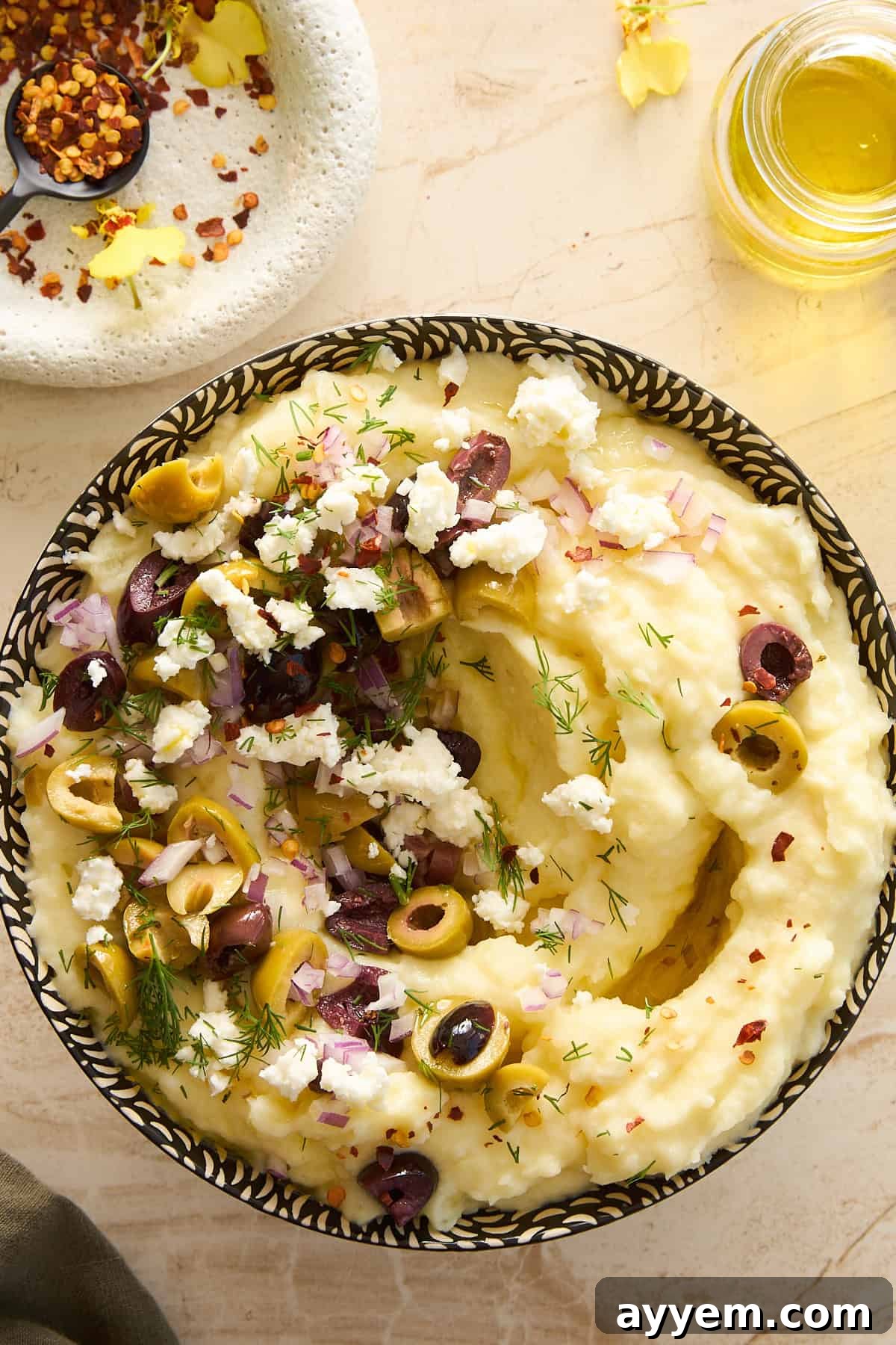 A bowl of creamy mashed potatoes topped with chopped olives, crumbled feta cheese, diced red onions, and fresh dill, with a drizzle of olive oil. A small dish of red pepper flakes and a jar of olive oil are nearby.