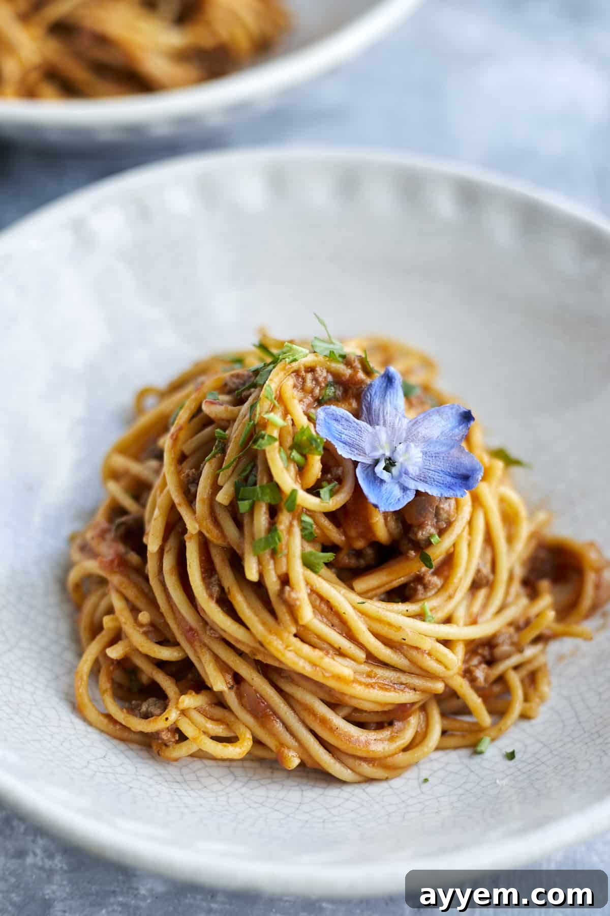 A close-up image of a white plate holding a generous serving of one-pot spaghetti with ground beef, beautifully topped with fresh herbs and a decorative blue flower.