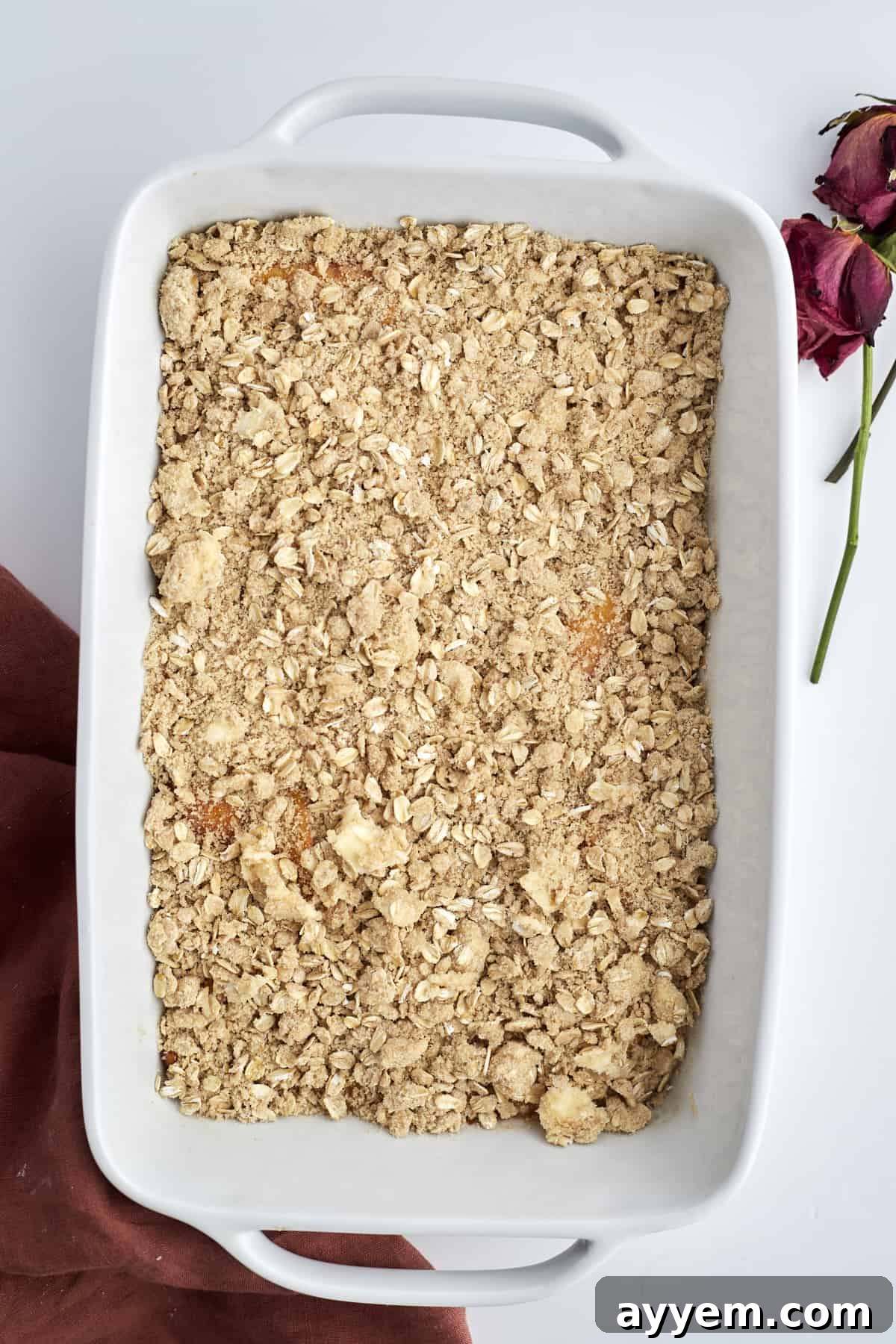 Delightfully Simple Peach Crisp 7 Overhead image of an unbaked peach crisp elegantly arranged in a white oval baking dish, showcasing the vibrant peach filling beneath a layer of golden, crumbly oat topping, ready for the oven.
