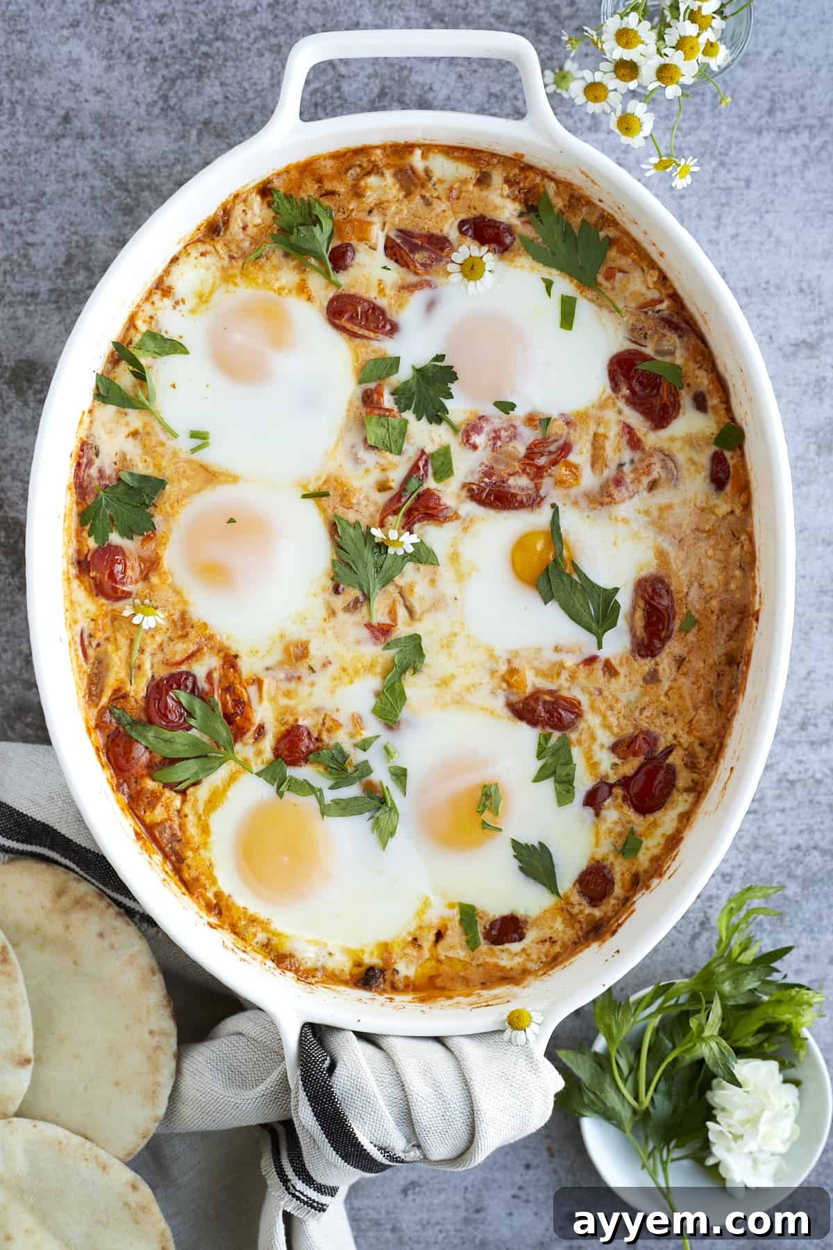 overhead image of 6 oven baked eggs in a white baking dish surrounding by vegetables and melted feta cheese topped with fresh herbs.