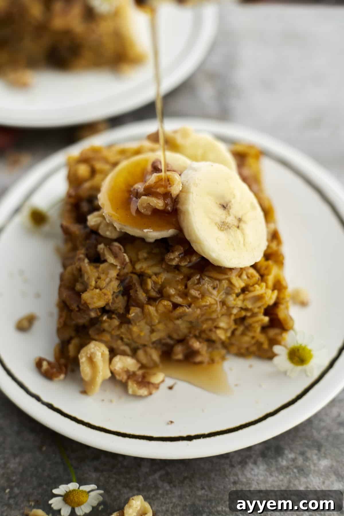A slice of baked pumpkin oatmeal topped with walnuts and banana coins with a drizzle of syrup being poured on top.