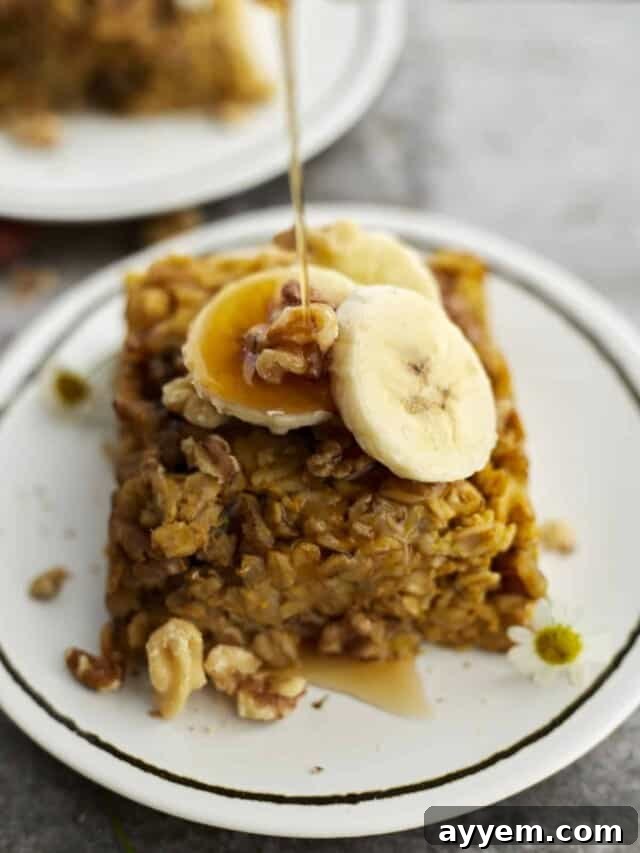A piece of baked pumpkin oatmeal on a white plate topped with walnuts and banana coins with a drizzle of maple syrup being poured on top.