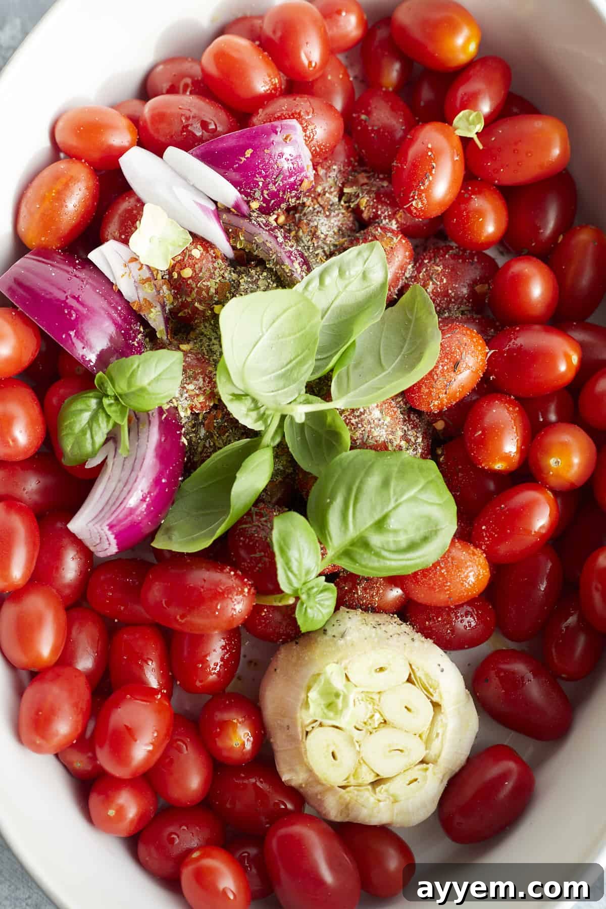 a baking dish with cherry tomatoes, a whole head of garlic, red onion, shallot, basil, and seasonings