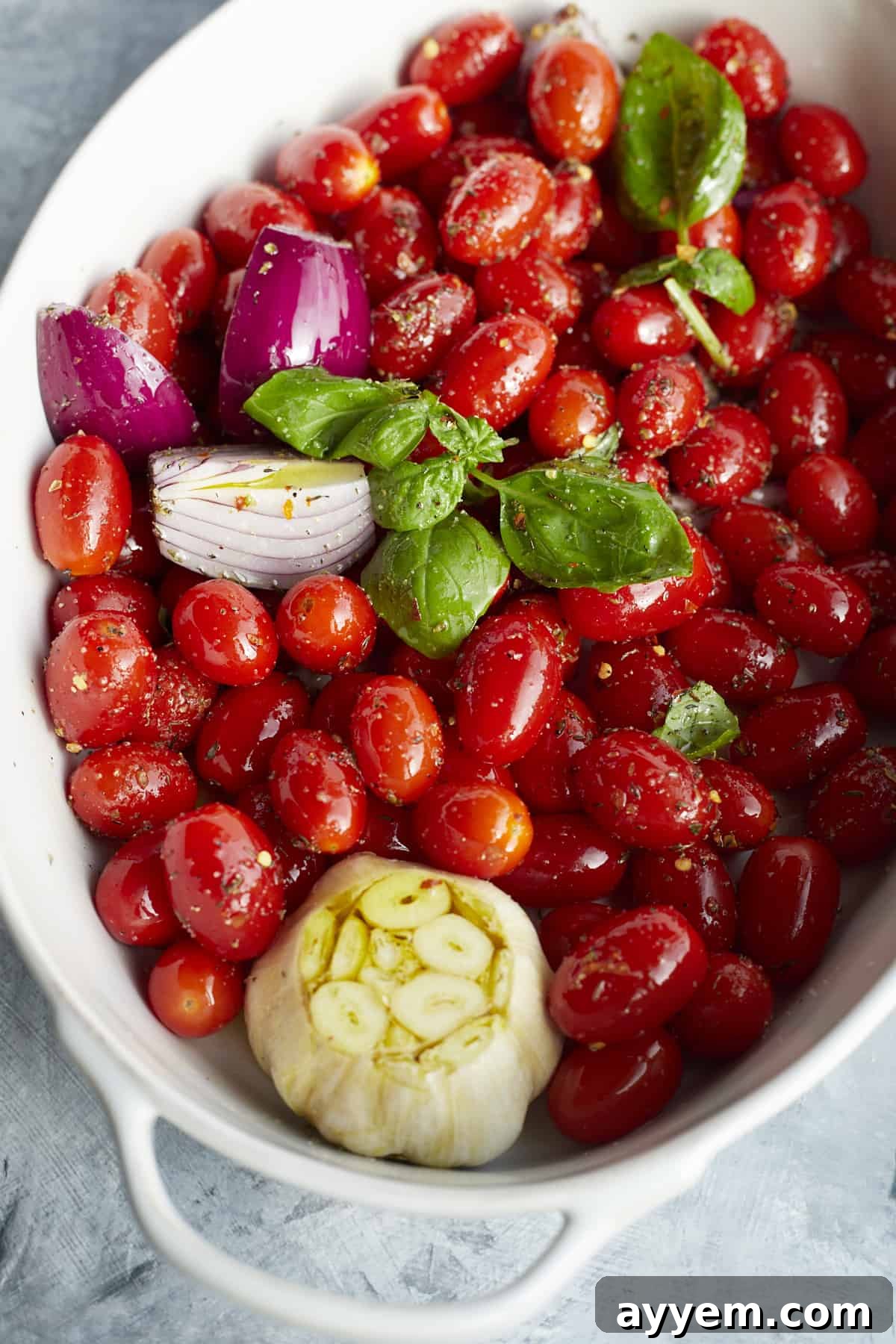 overhead image of tomatoes, garlics, shallot, onions, basil, and seasoning drizzled with olive oil in a white baking dish