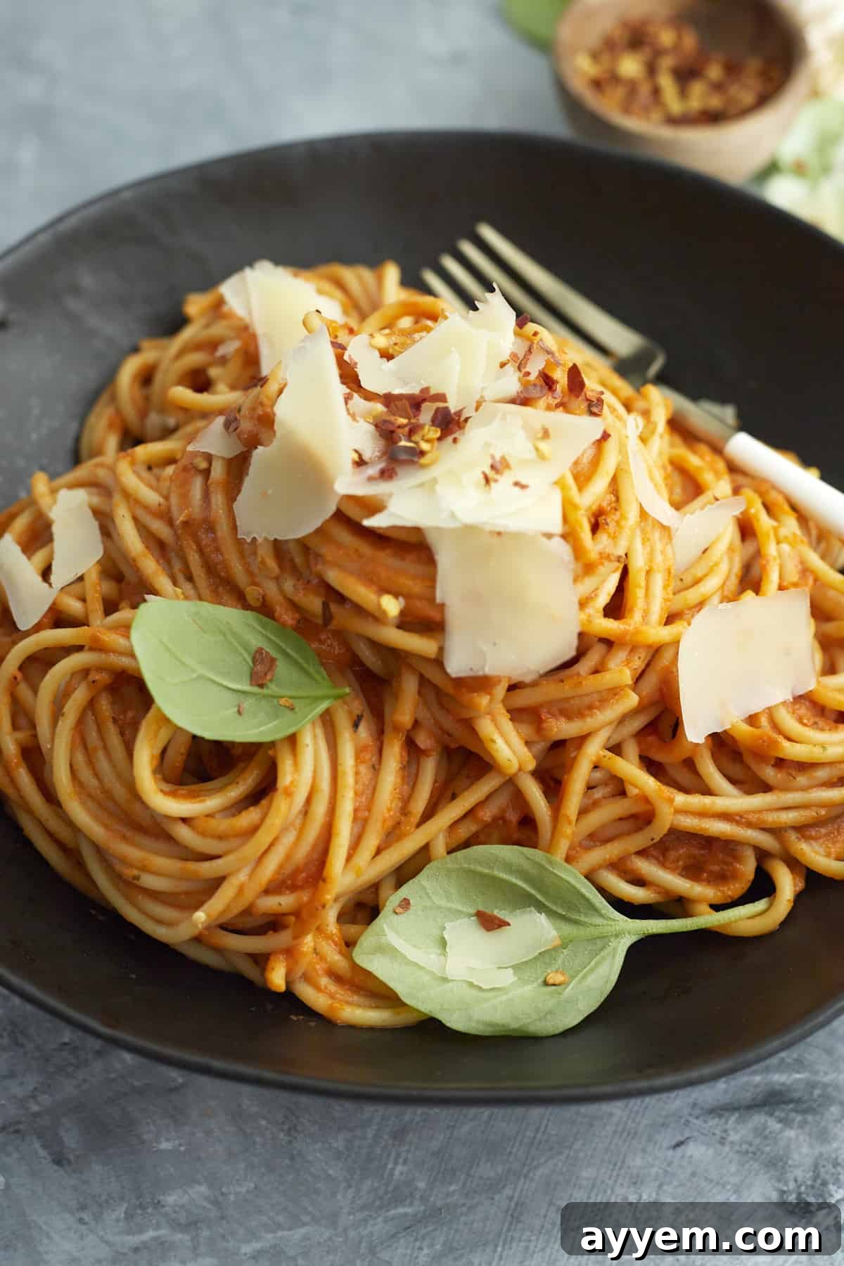 a black plate with a pile of arrabiata sauce spaghetti bake topped with Parmesan cheese, basil leaves, and red pepper flakes